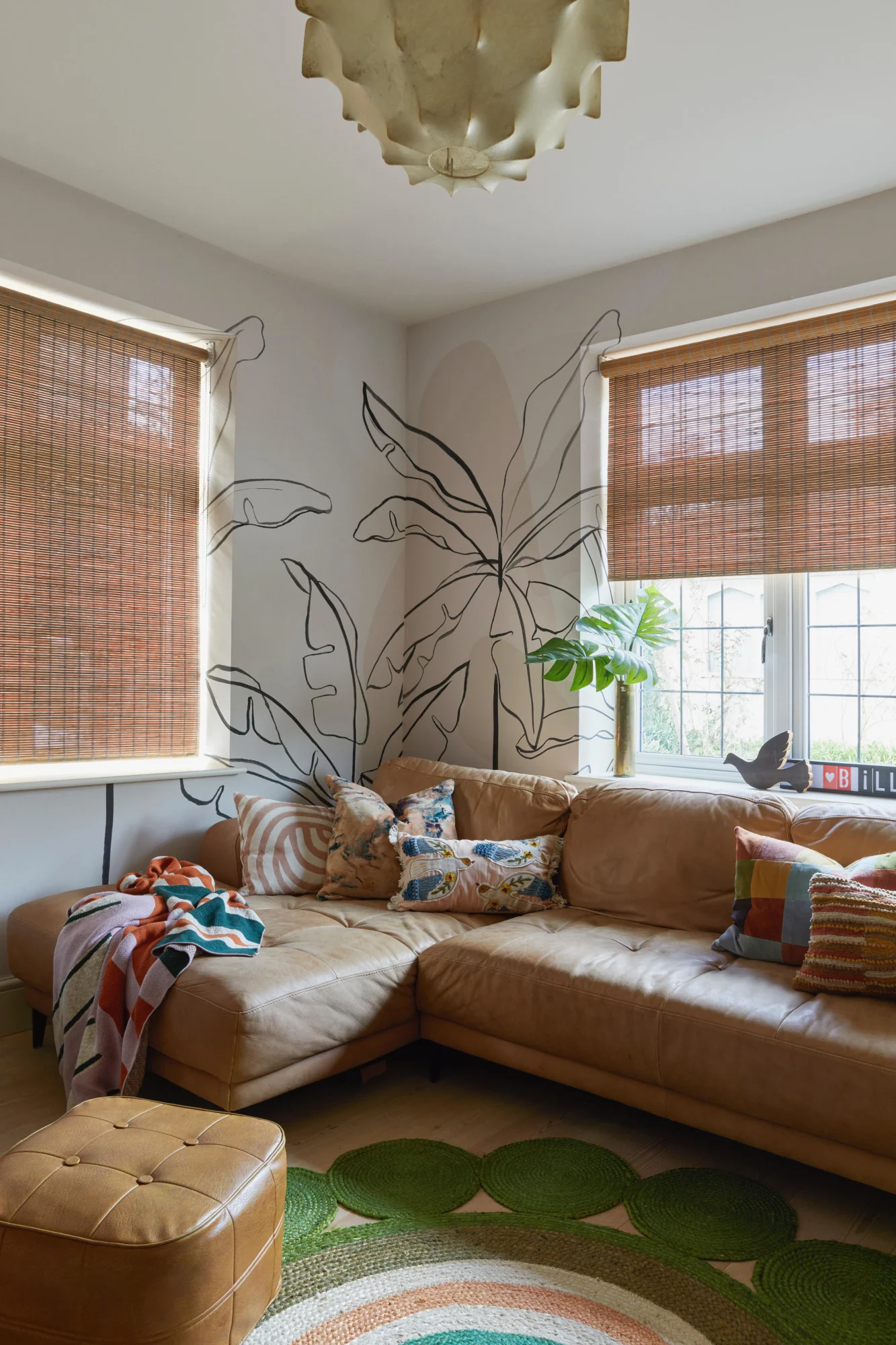 This living room features a large brown leather sectional sofa, 2 windows with wooden blinds, a palm tree wall mural, and colorful cushions and throws.
