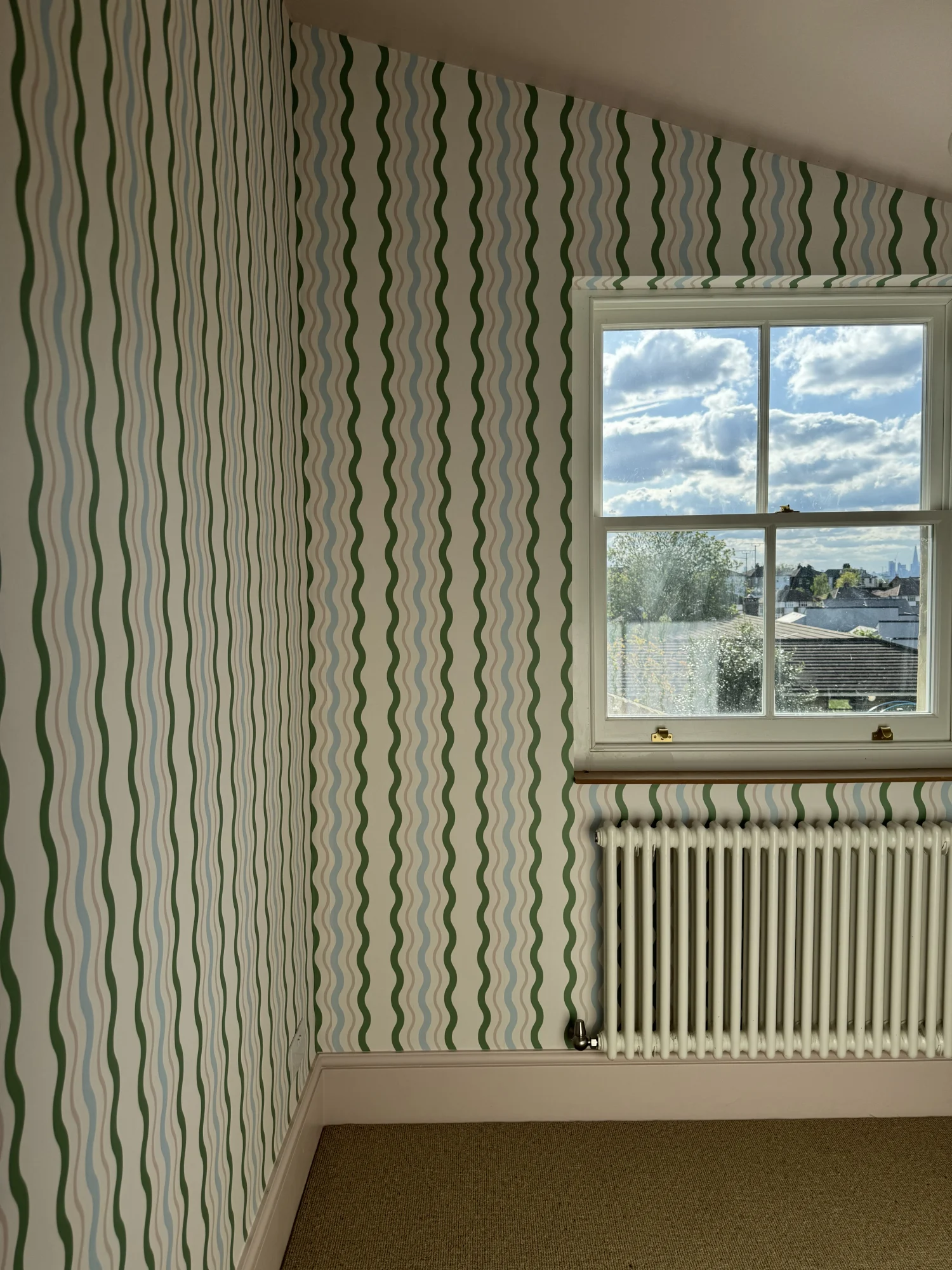 This room has wavy green and pink striped wallpaper, a white radiator, and a window overlooking a cloudy sky.