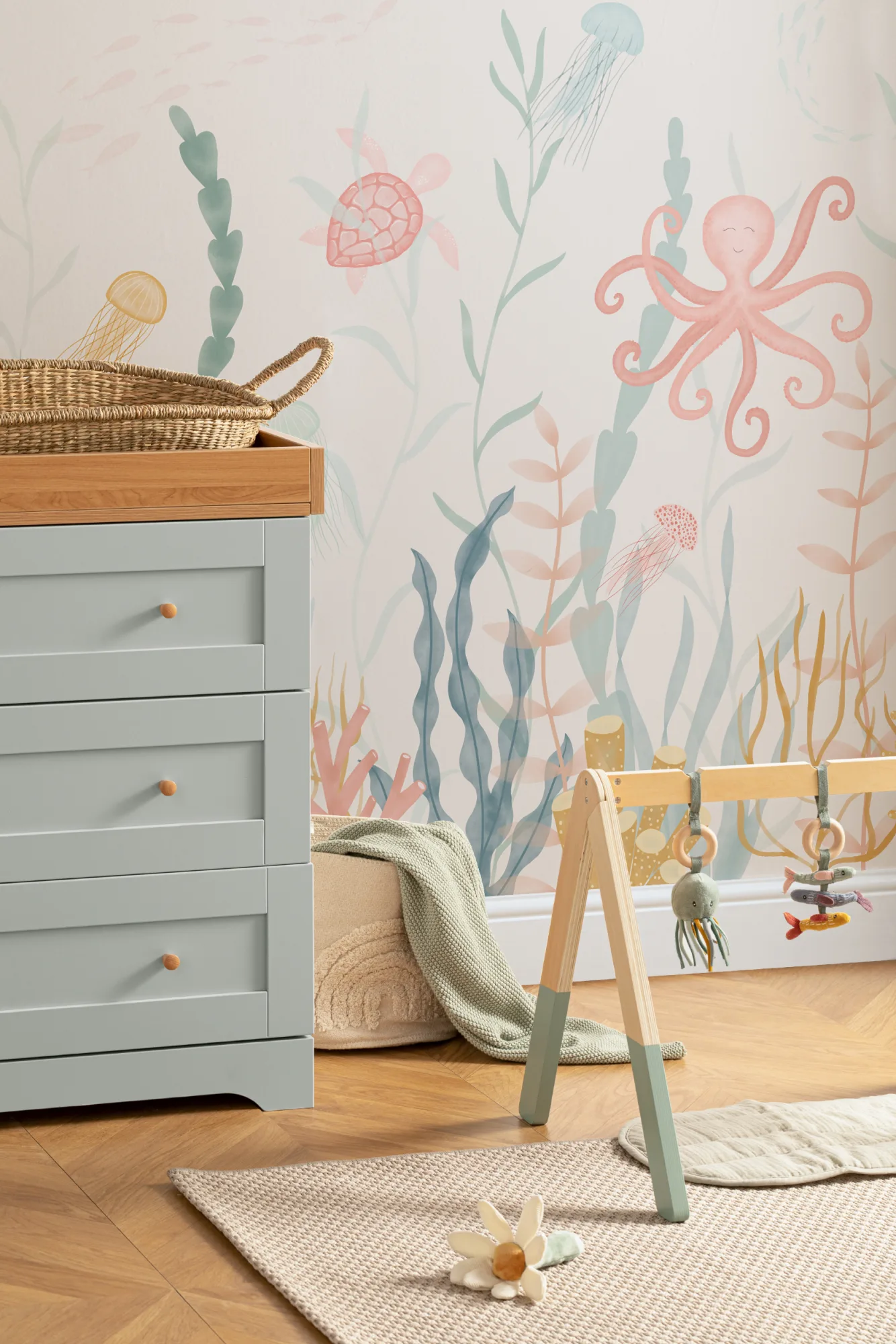 An undersea-themed wall mural with pastel sea creatures in a nursery with a green dresser and natural woven basket.