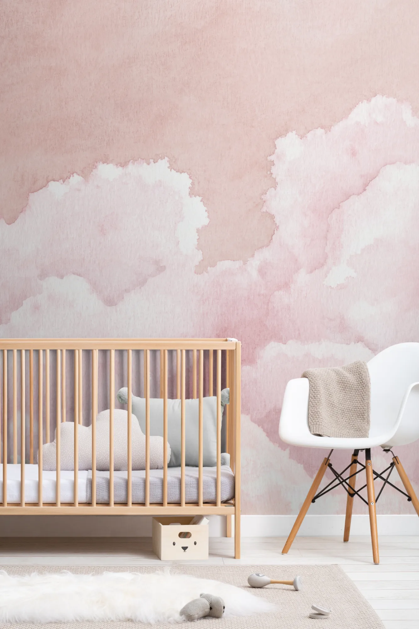 A soft pink nursery with a cloud mural, wooden crib, and white modern chair.