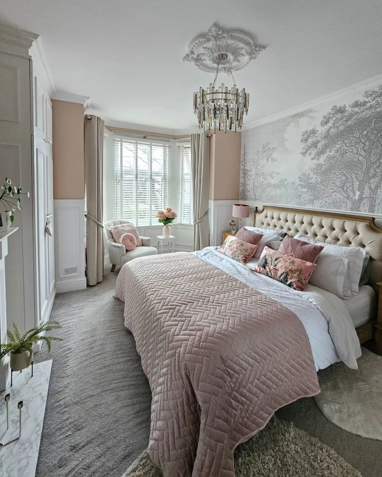 A serene bedroom with a neutral-toned forest wall mural, tufted headboard, soft pink bedding, and a chic chandelier.
