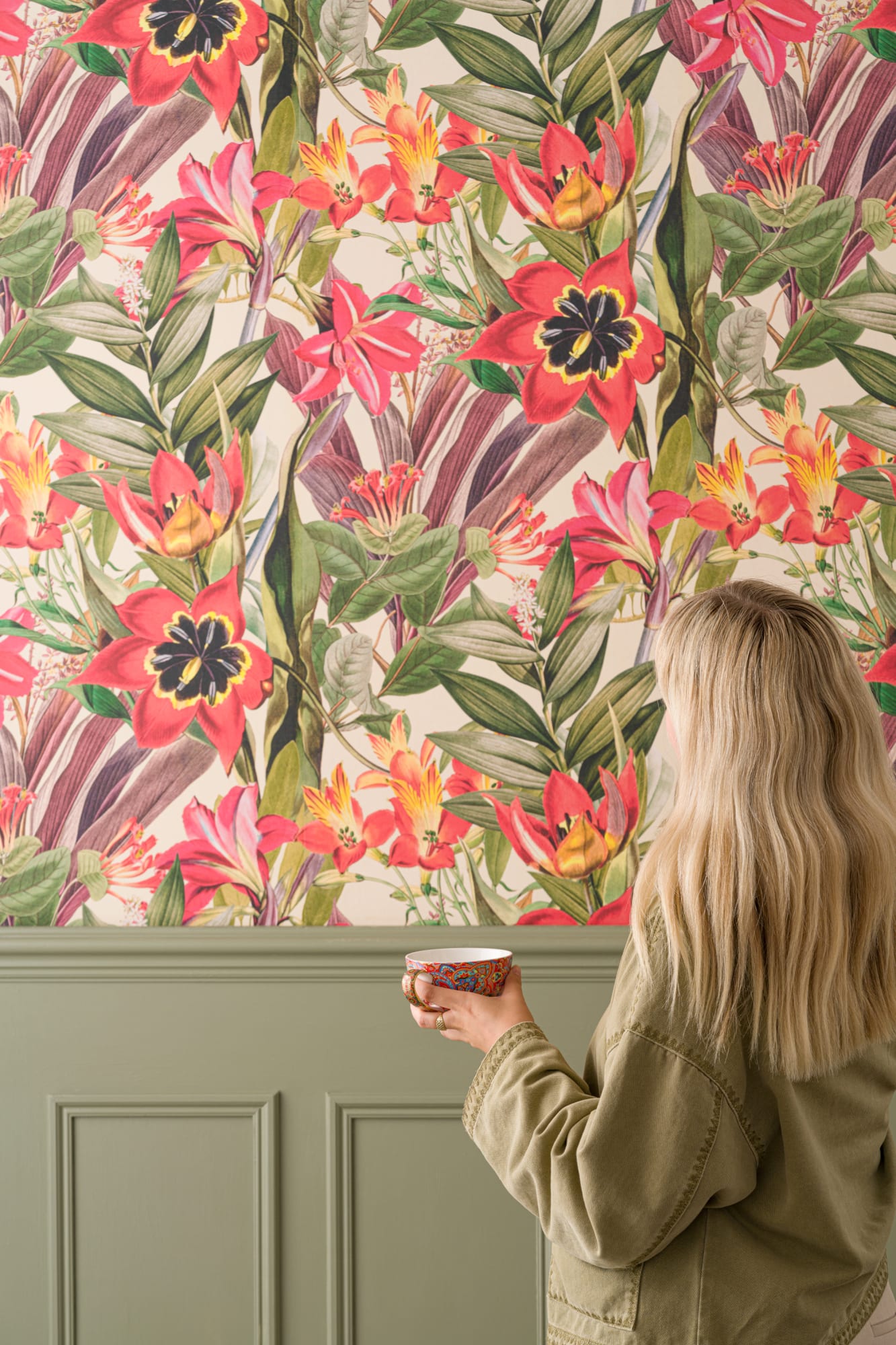 A vibrant wall featuring tropical floral wallpaper with red, pink, and yellow flowers, paired with green wainscoting panelling and a woman holding a patterned mug.