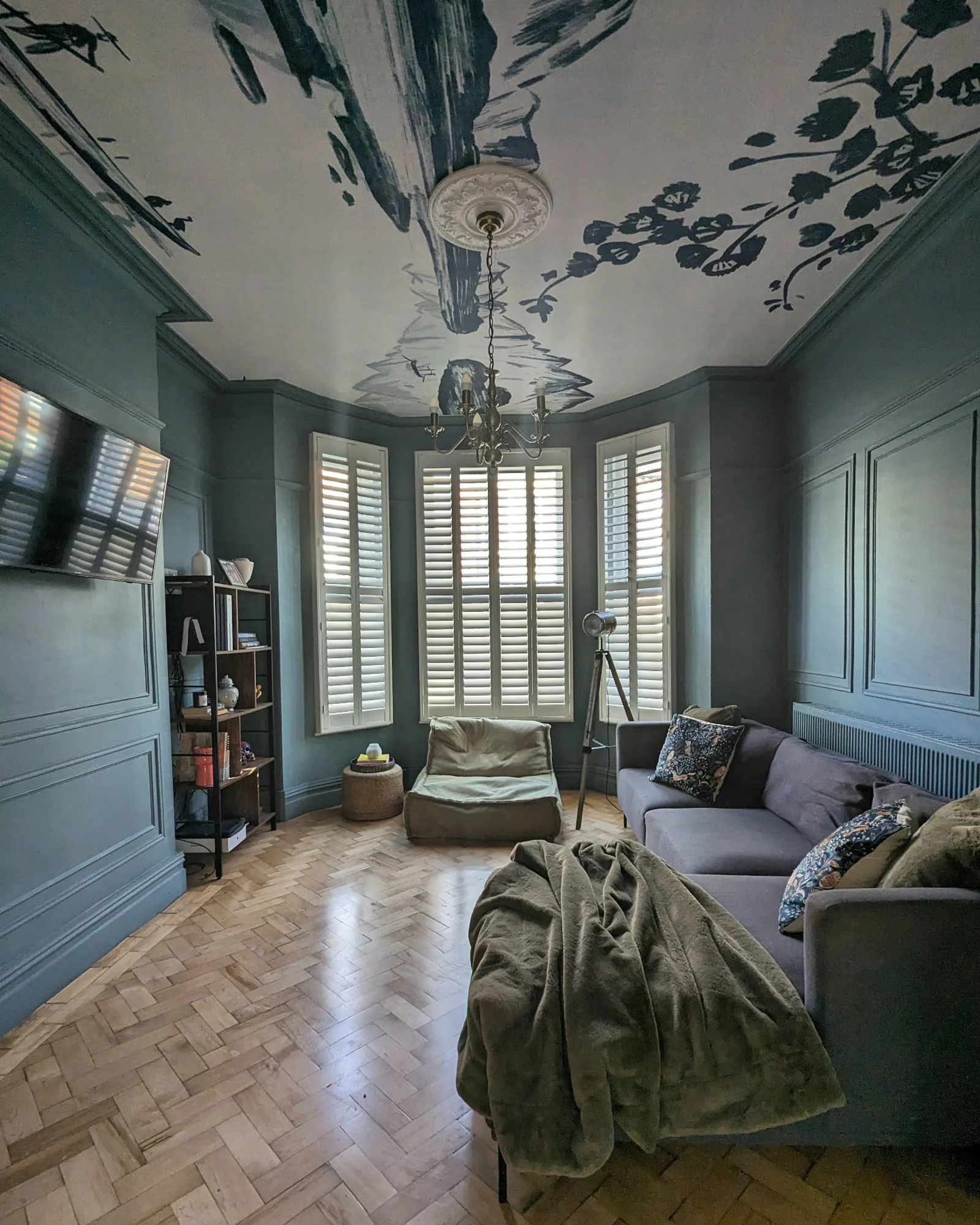 A cozy living room with deep blue walls and ceiling, featuring a painted mural, parquet flooring, and elegant shutters framing the windows.
