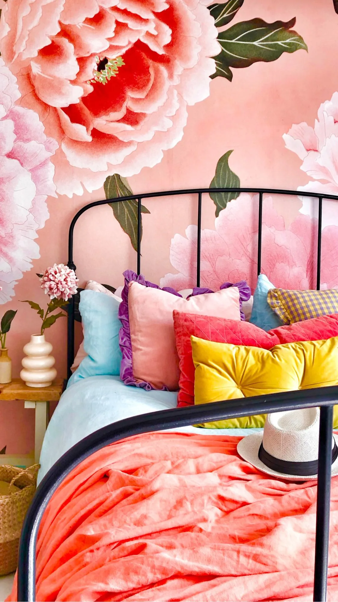 A bright bedroom with oversized pink peony wallpaper, a black metal bedframe, and colorful pillows in vibrant hues.