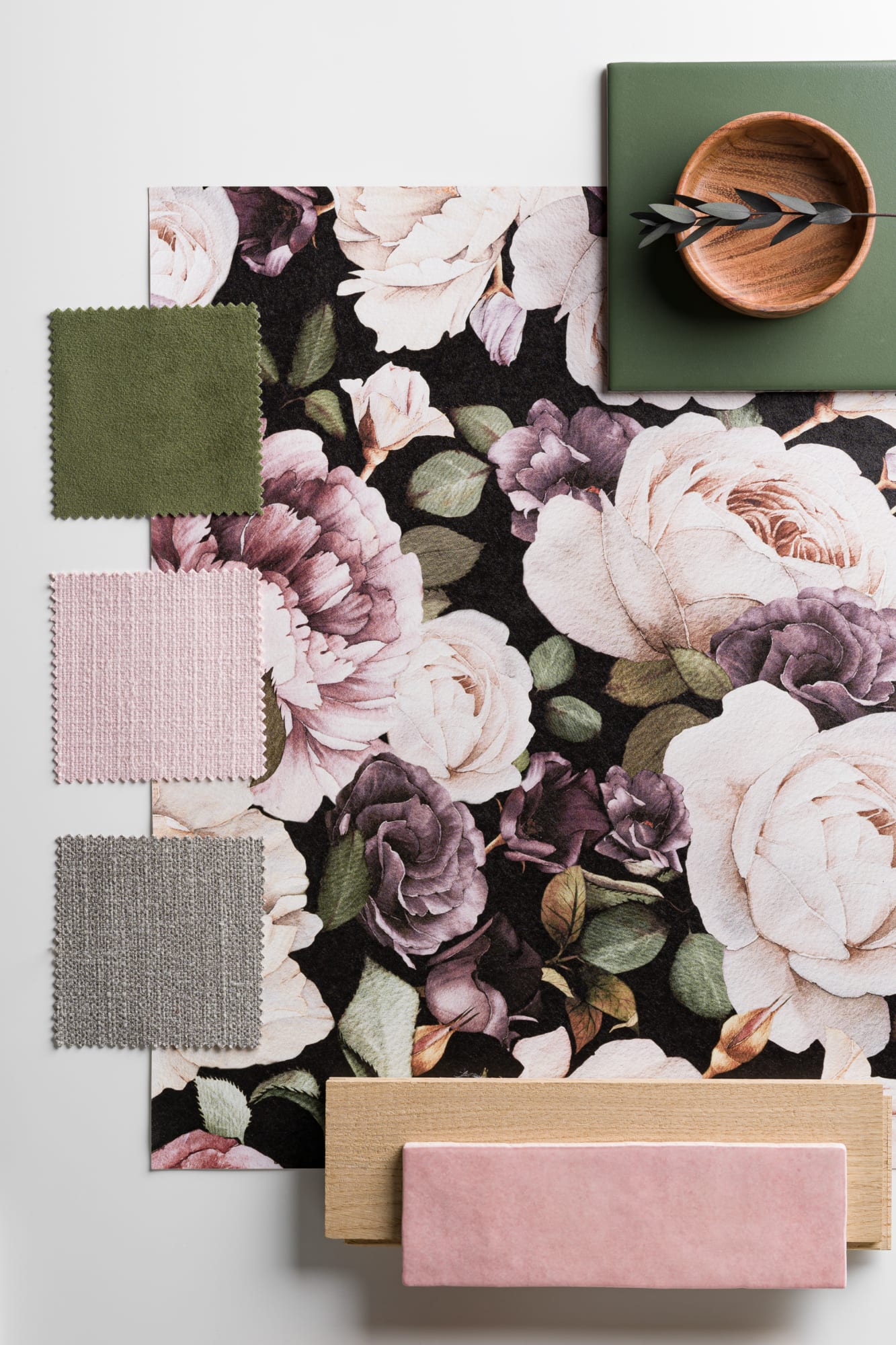A styled mood board showcasing a bold floral wallpaper sample alongside fabric swatches in pink, green, and neutral tones, with wood and ceramic accents.