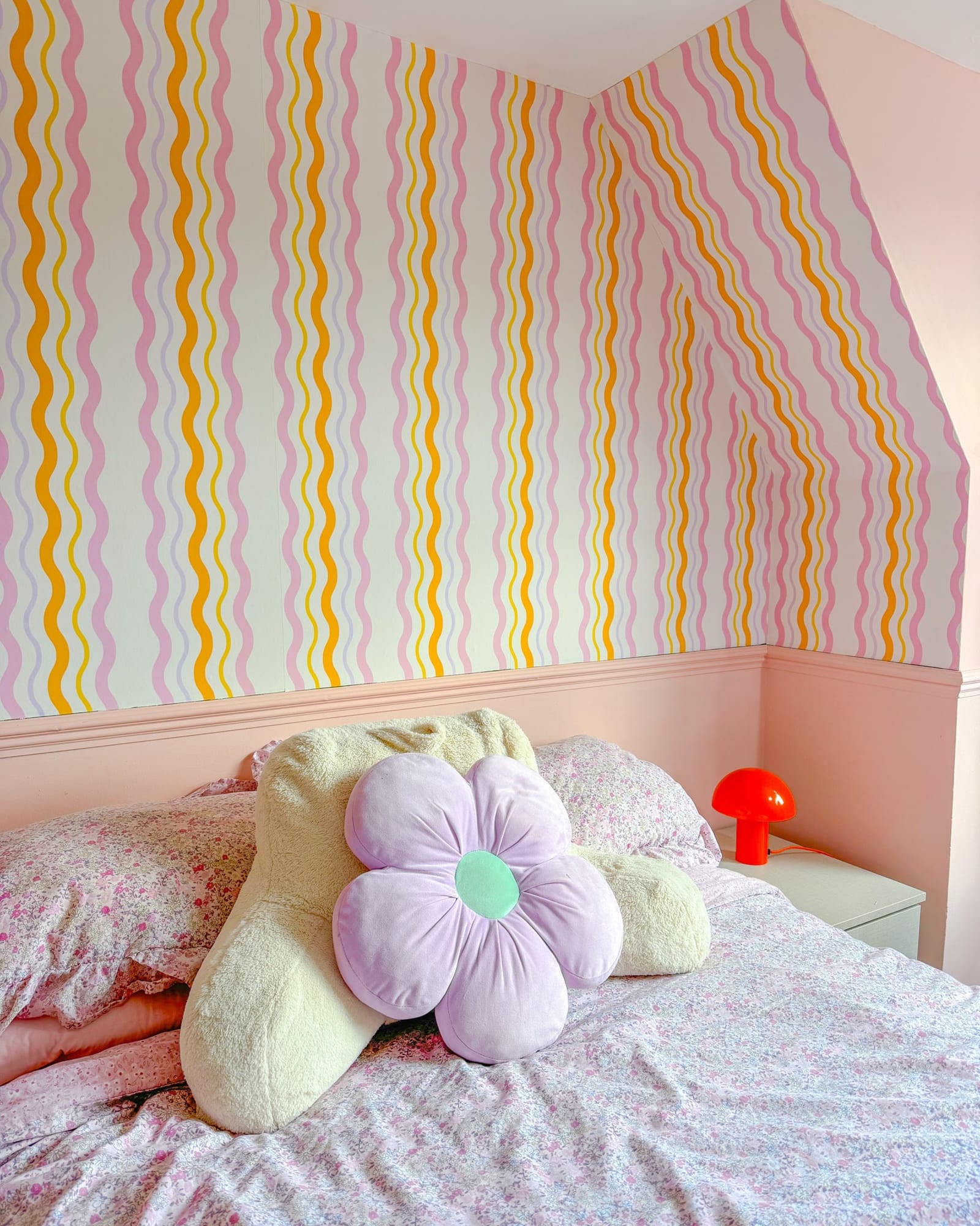 A cozy bed with floral bedding and a pastel wavy striped wallpaper on an angled wall.
