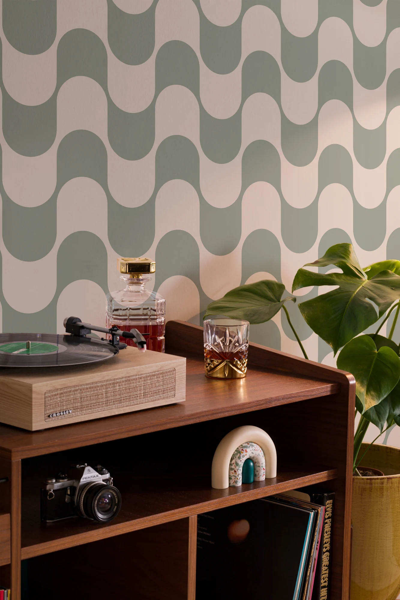 A record player and decor sit against a backdrop of green wavy patterned wallpaper with a mid-century modern vibe.
