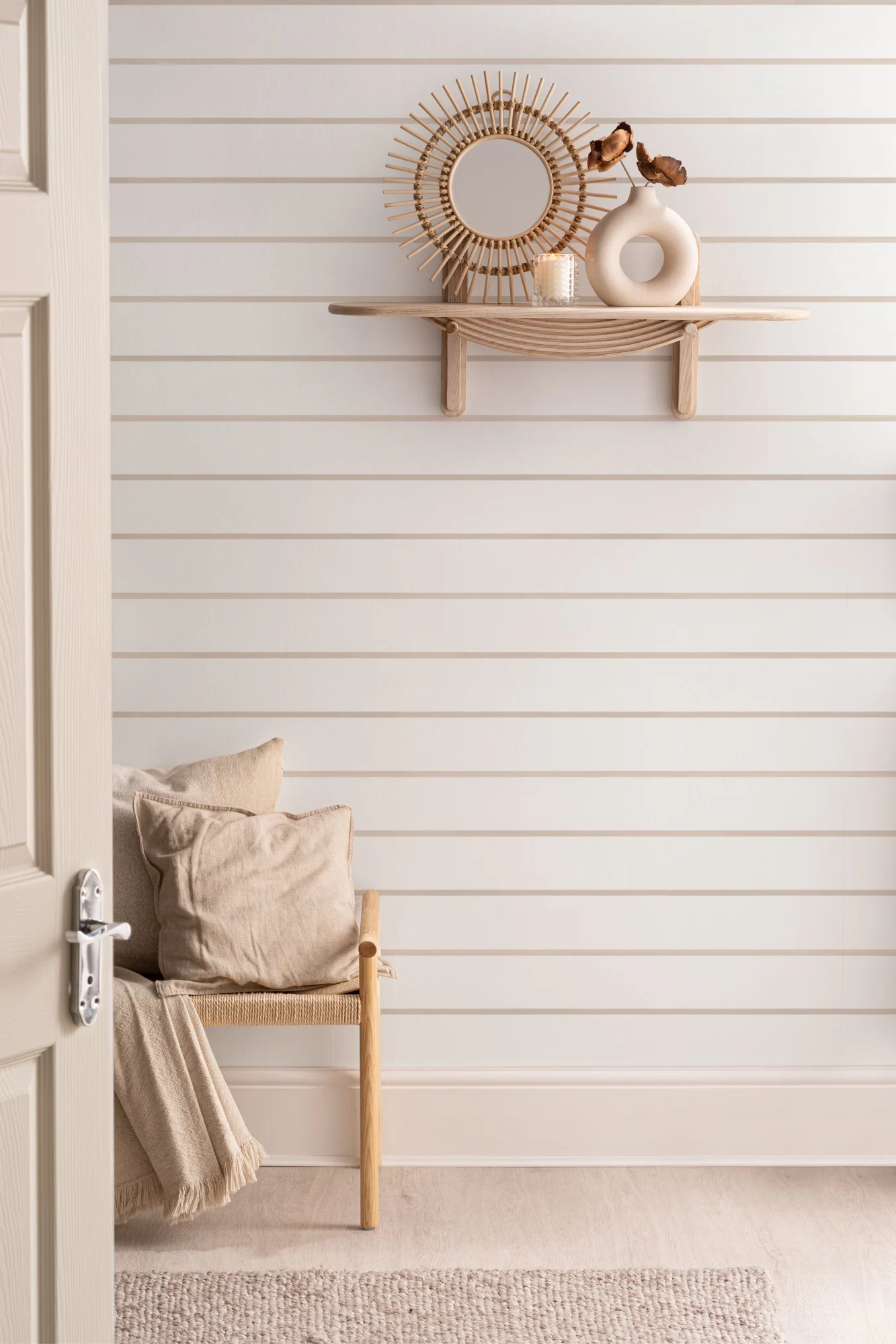 An airy Scandi style hallway with horizontal striped wallpaper, a wooden shelf holding decor, and a woven chair with a cozy throw.