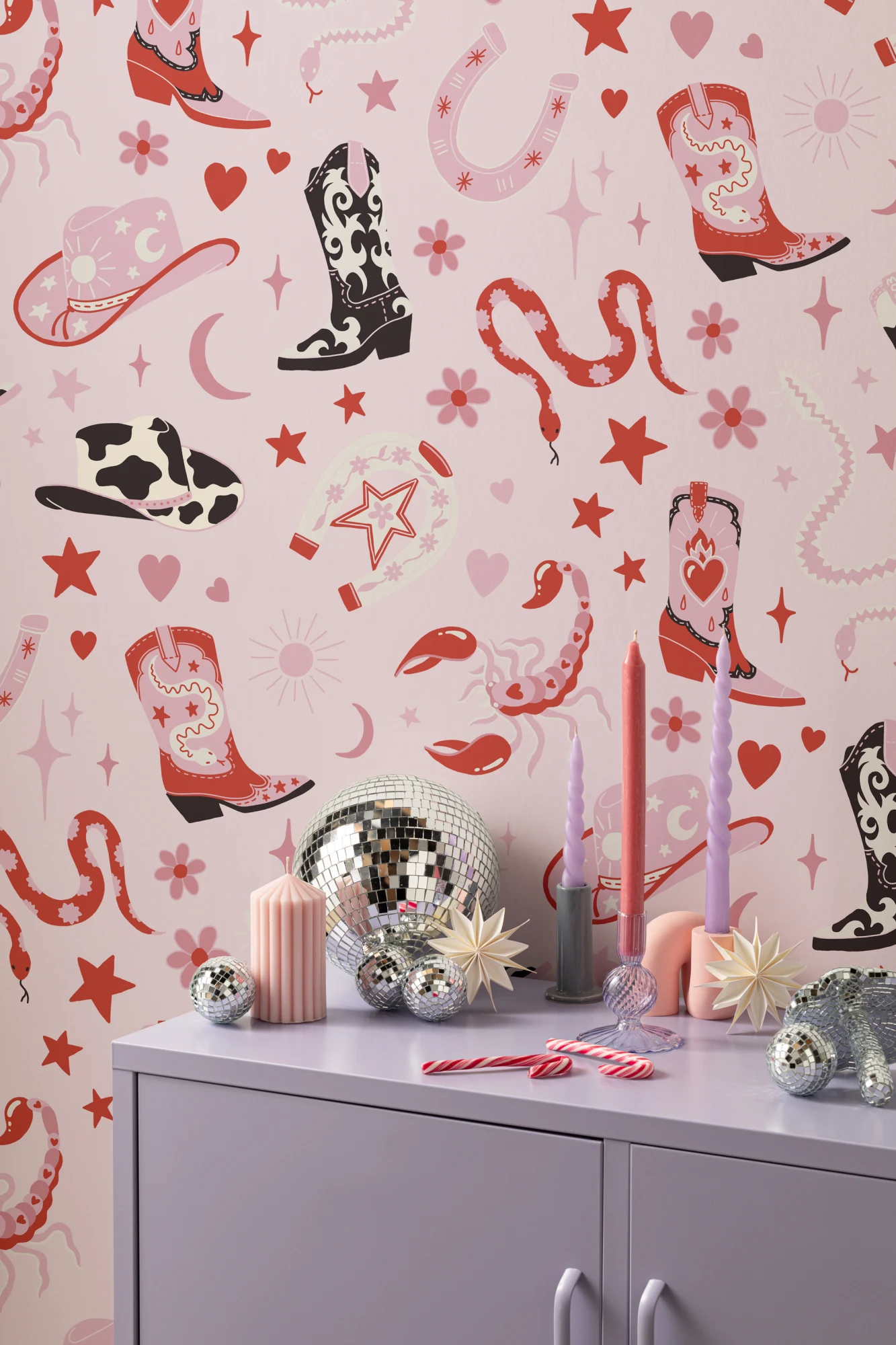 A playful cowboy-themed maximalist wallpaper with illustrations of boots, hats, snakes, and stars in shades of pink and red, styled with disco balls and pastel candles.