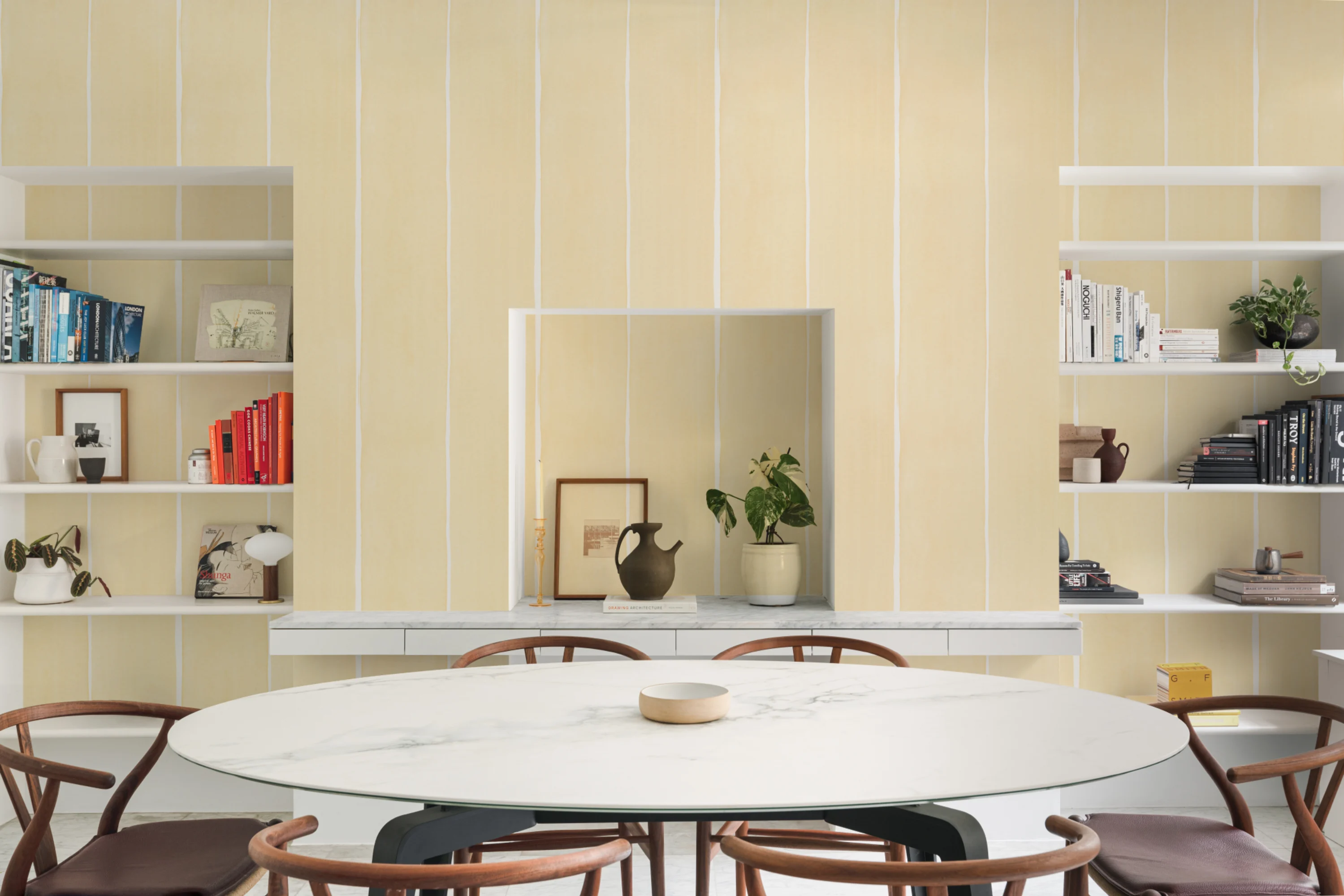 A modern dining room featuring soft yellow striped wallpaper, built-in shelves, and a white marble table.
