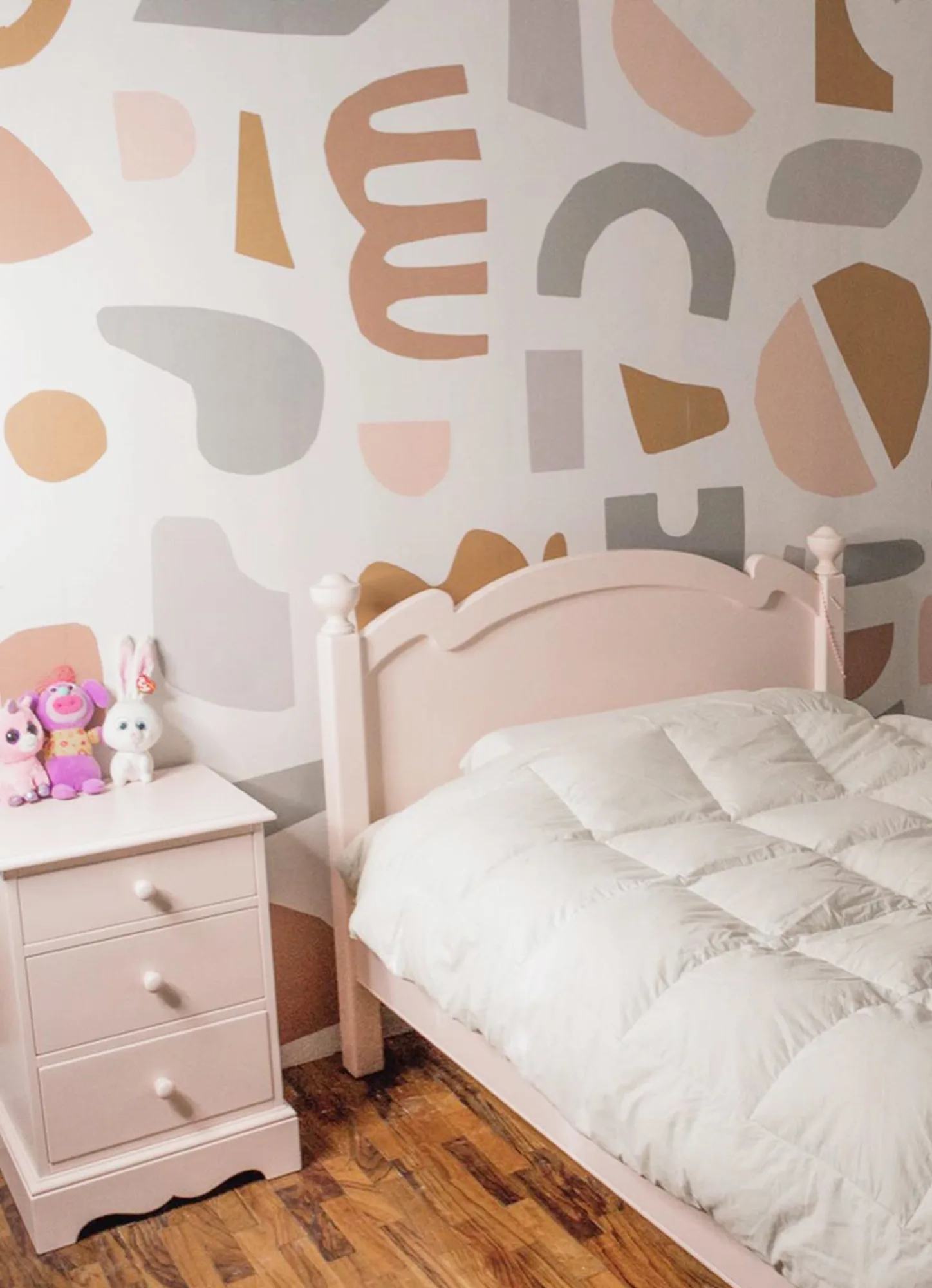 A playful kids' bedroom with a pastel abstract wallpaper, a pink bed, and a matching nightstand with stuffed animals.