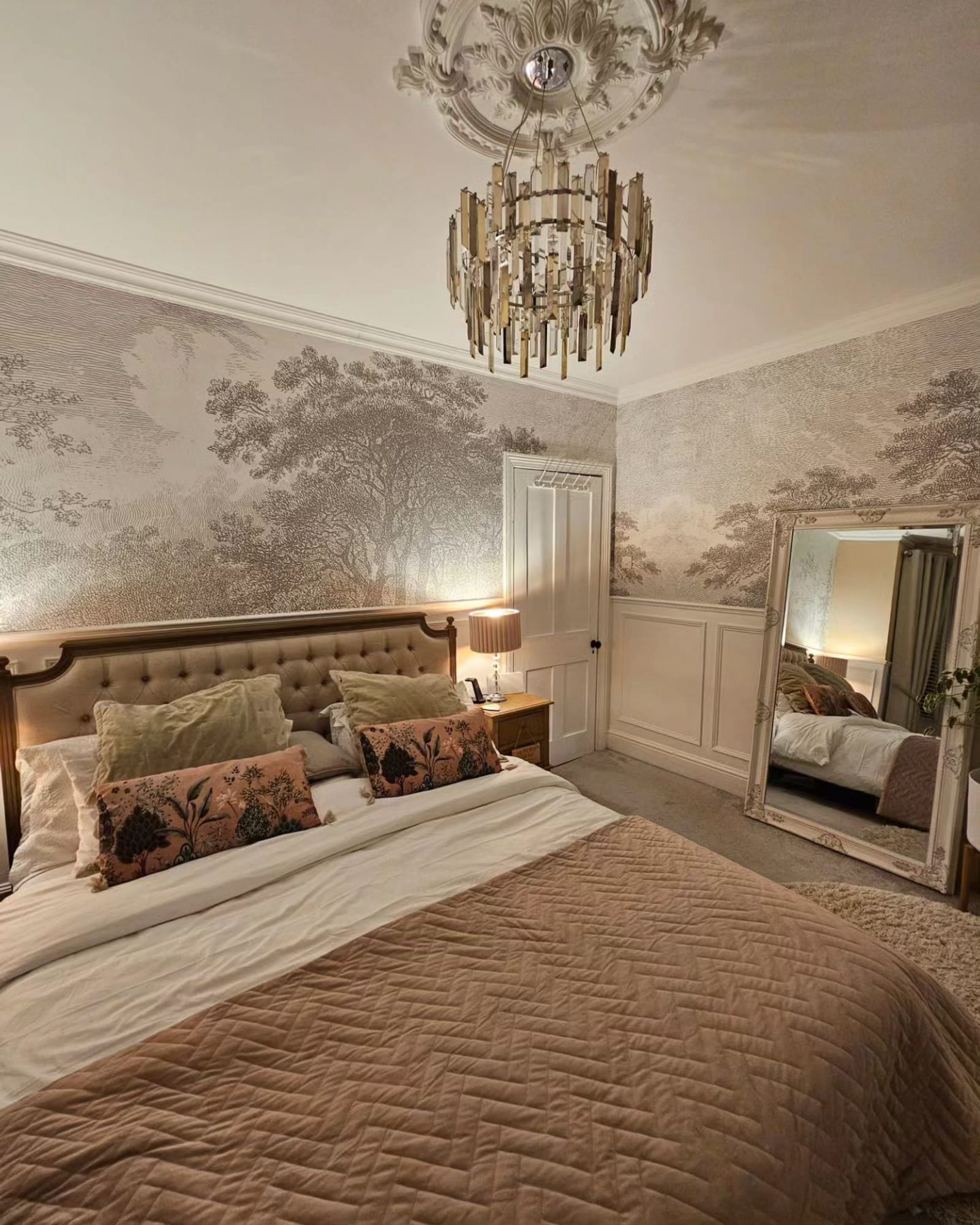 Cozy bedroom with detailed forest mural, tufted headboard, and a decorative ceiling chandelier.