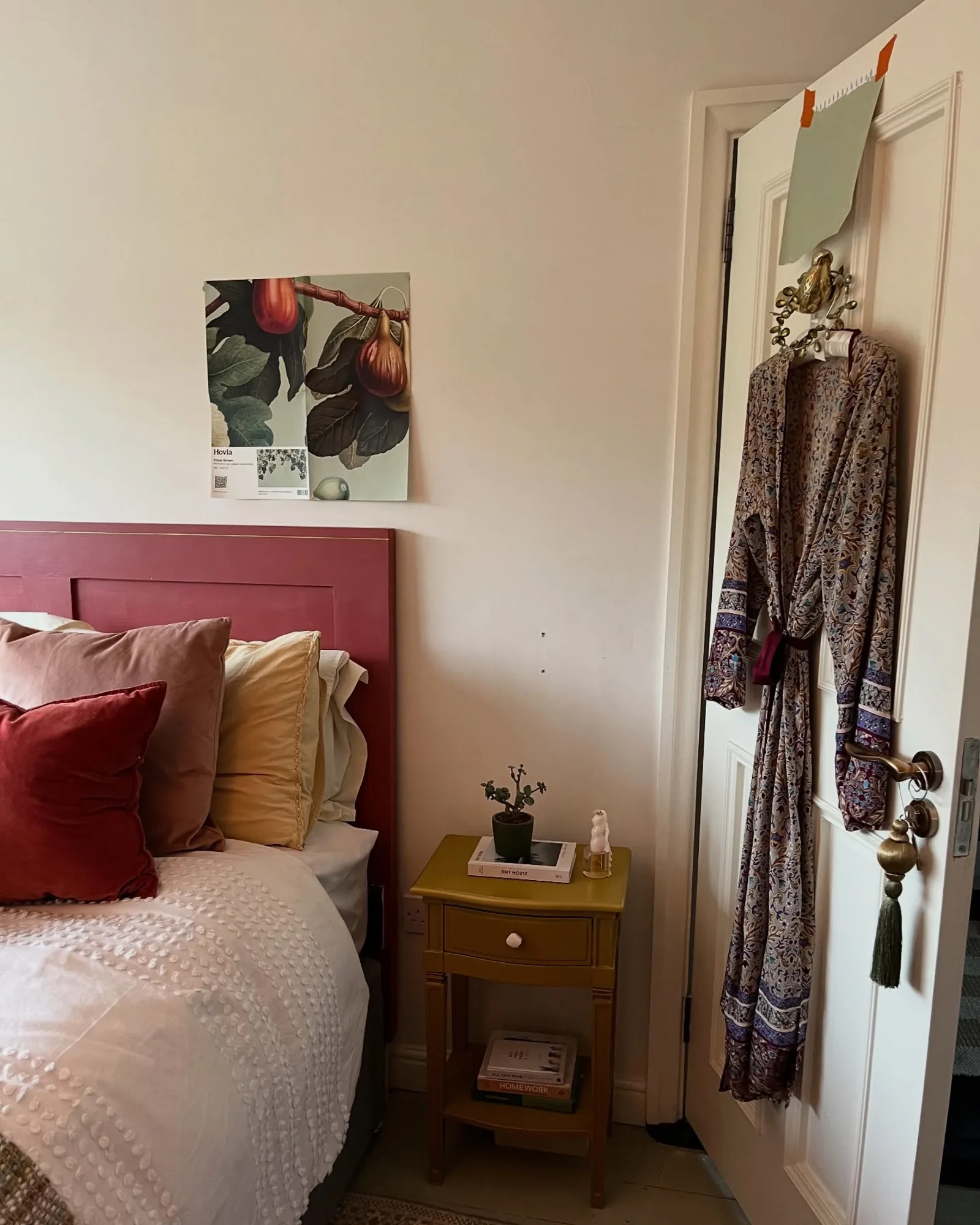 A bedroom corner with a red headboard, layered pillows, a mustard nightstand, and a botanical wallpaper sample taped to the wall.