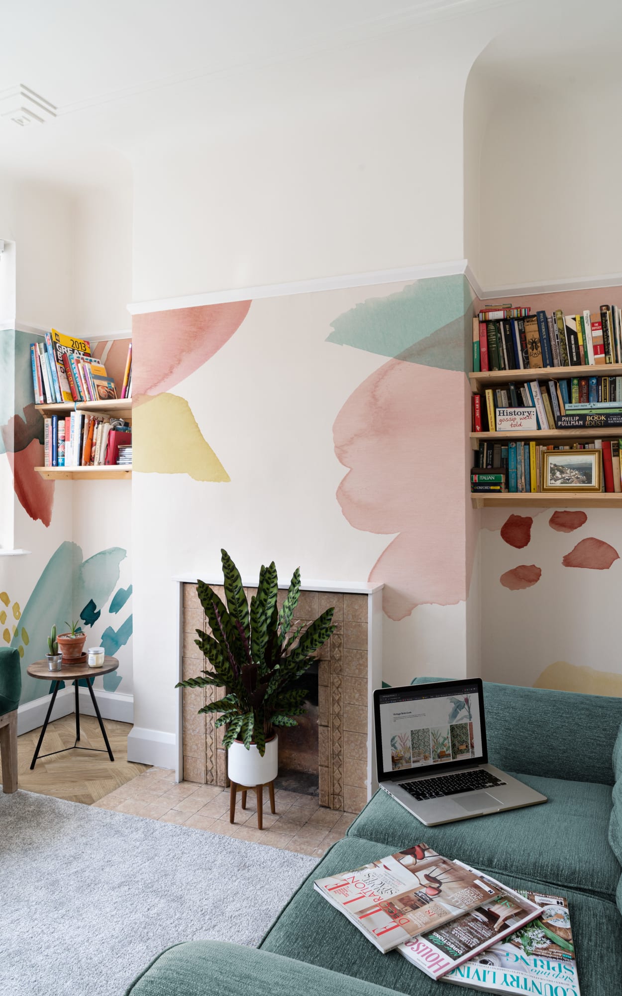 A light-filled living room with an abstract pastel mural surrounding a fireplace and bookshelves.