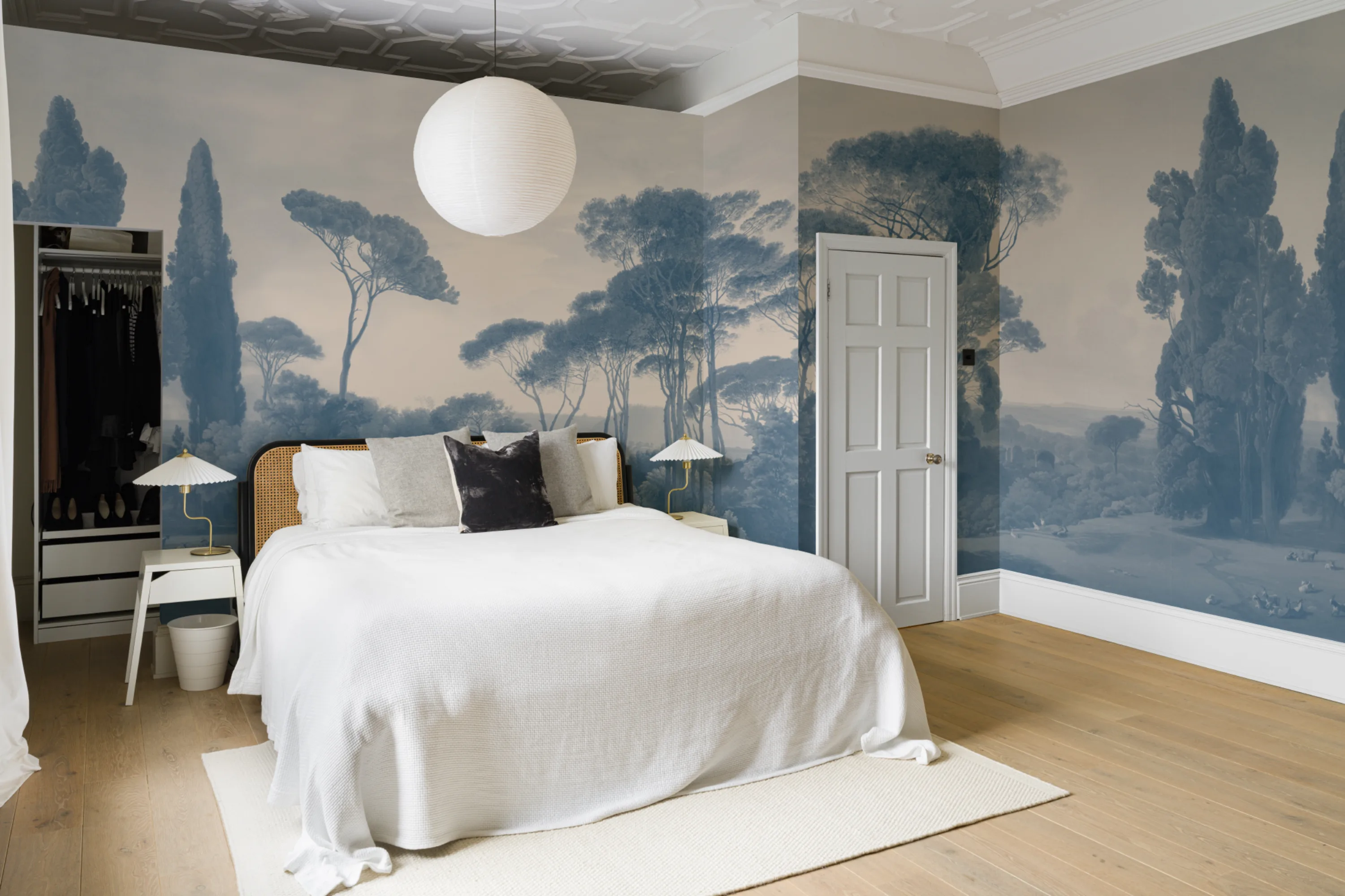 Serene bedroom with white bedding and blue-toned scenic mural of trees and hills wrapping around the walls.