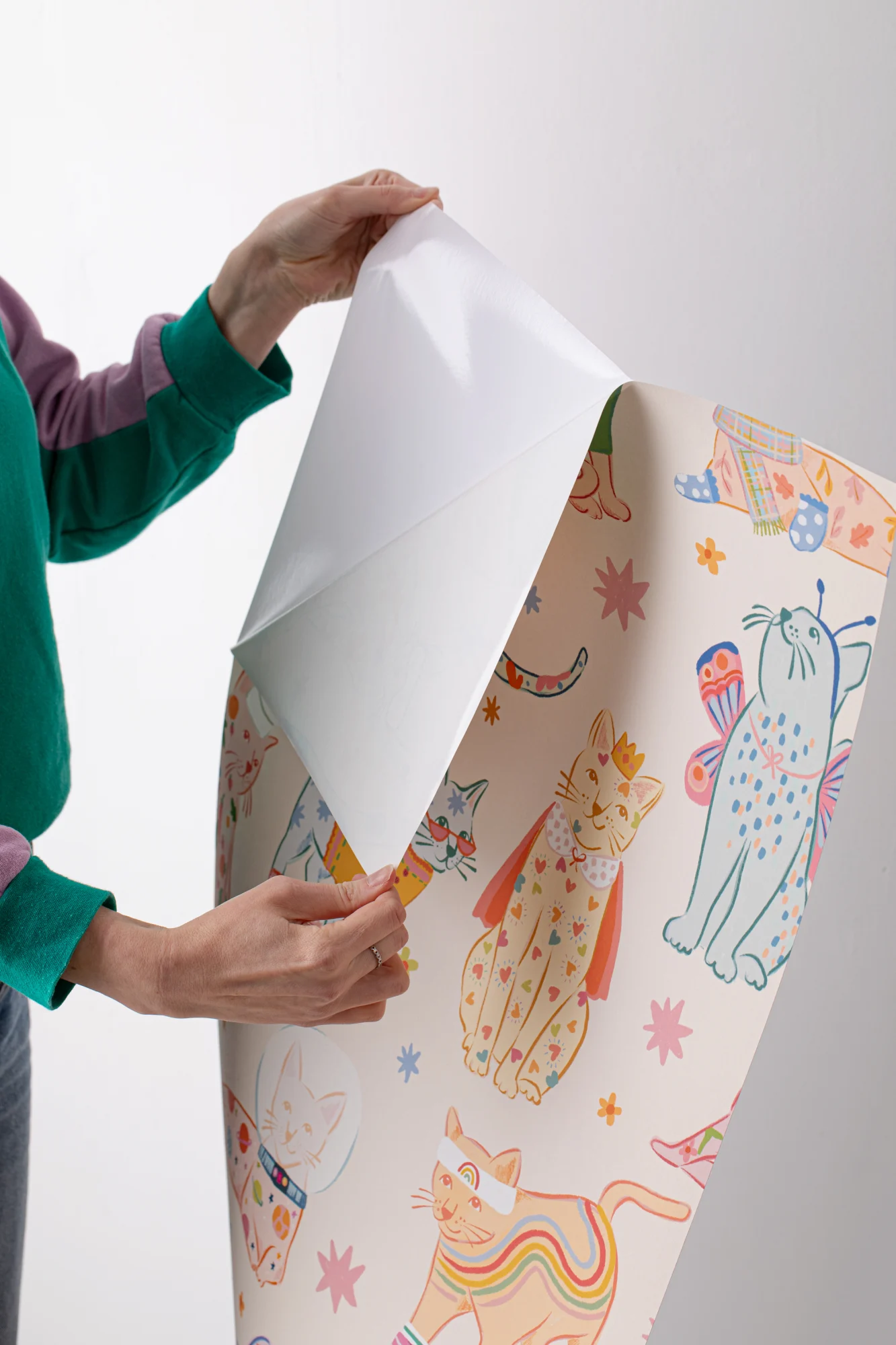 A person holds and peels a large sheet of illustrated peel-and-stick wallpaper featuring whimsical cats.