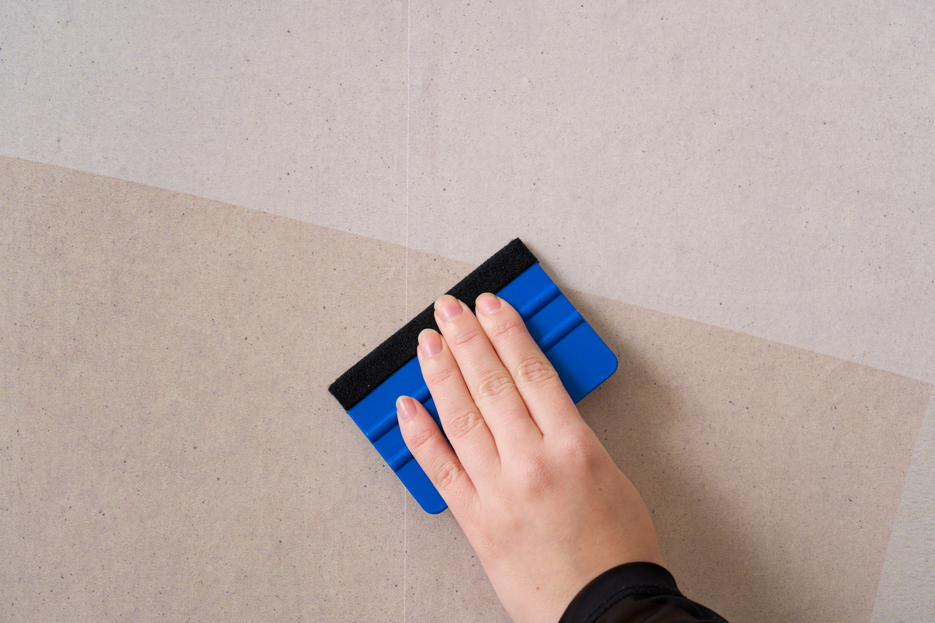 A hand uses a blue smoothing tool to press down peel-and-stick wallpaper on a wall.