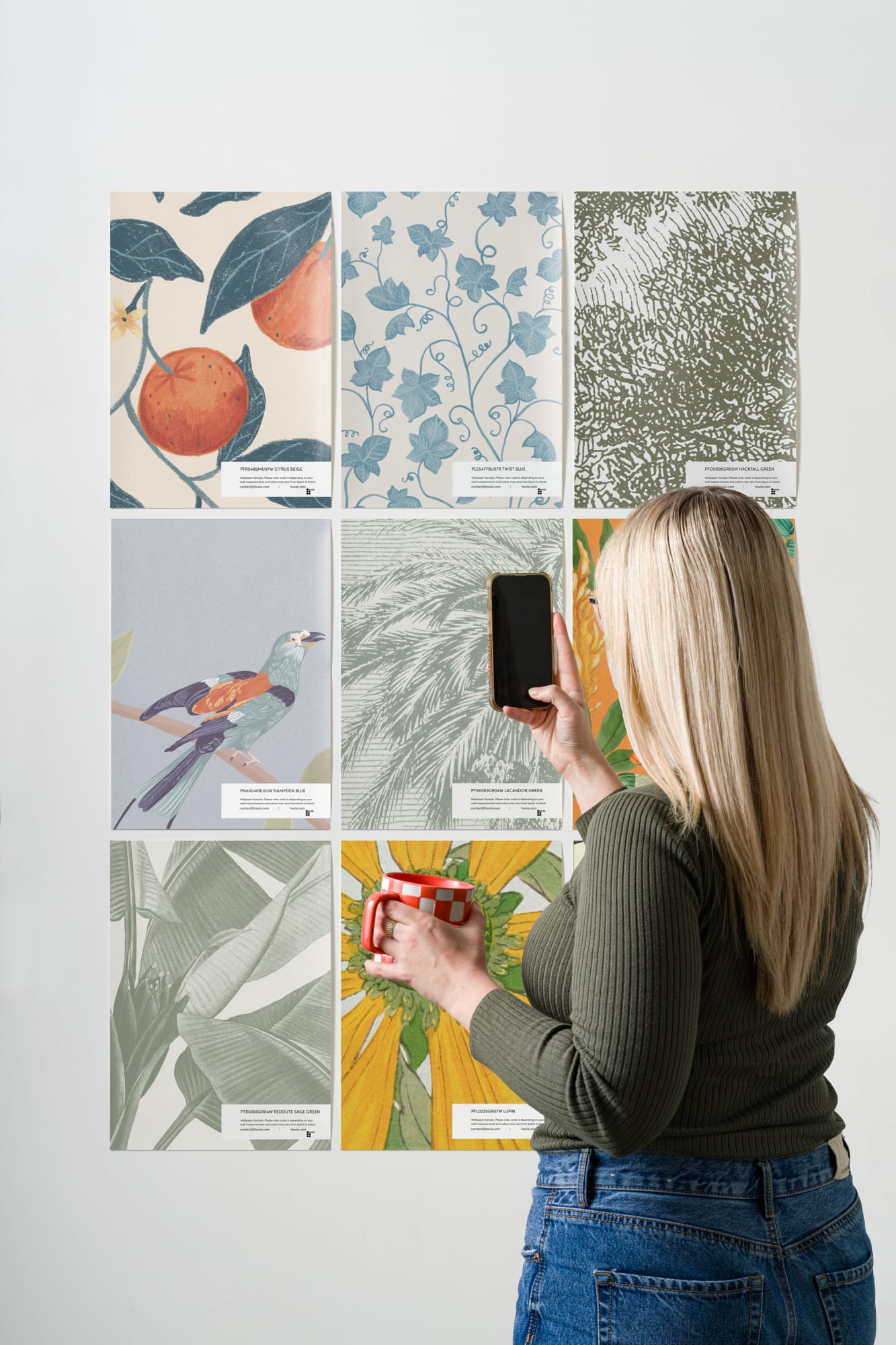 A woman with long blonde hair photographs a grid of botanical wallpaper samples on a white wall while holding a red-and-white mug.