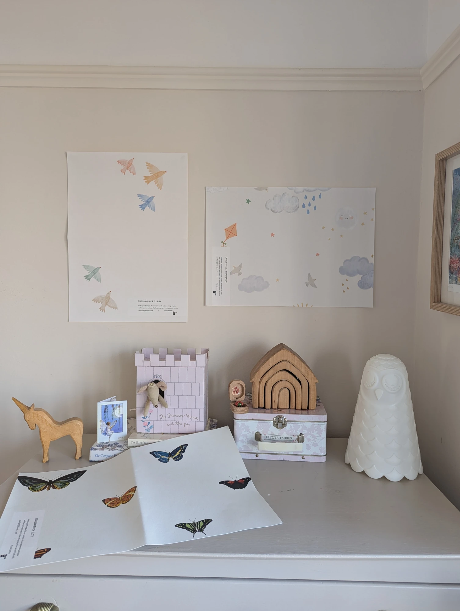 A collection of children’s wallpaper samples featuring birds, clouds, kites, and butterflies displayed above a dresser with toys and decor.