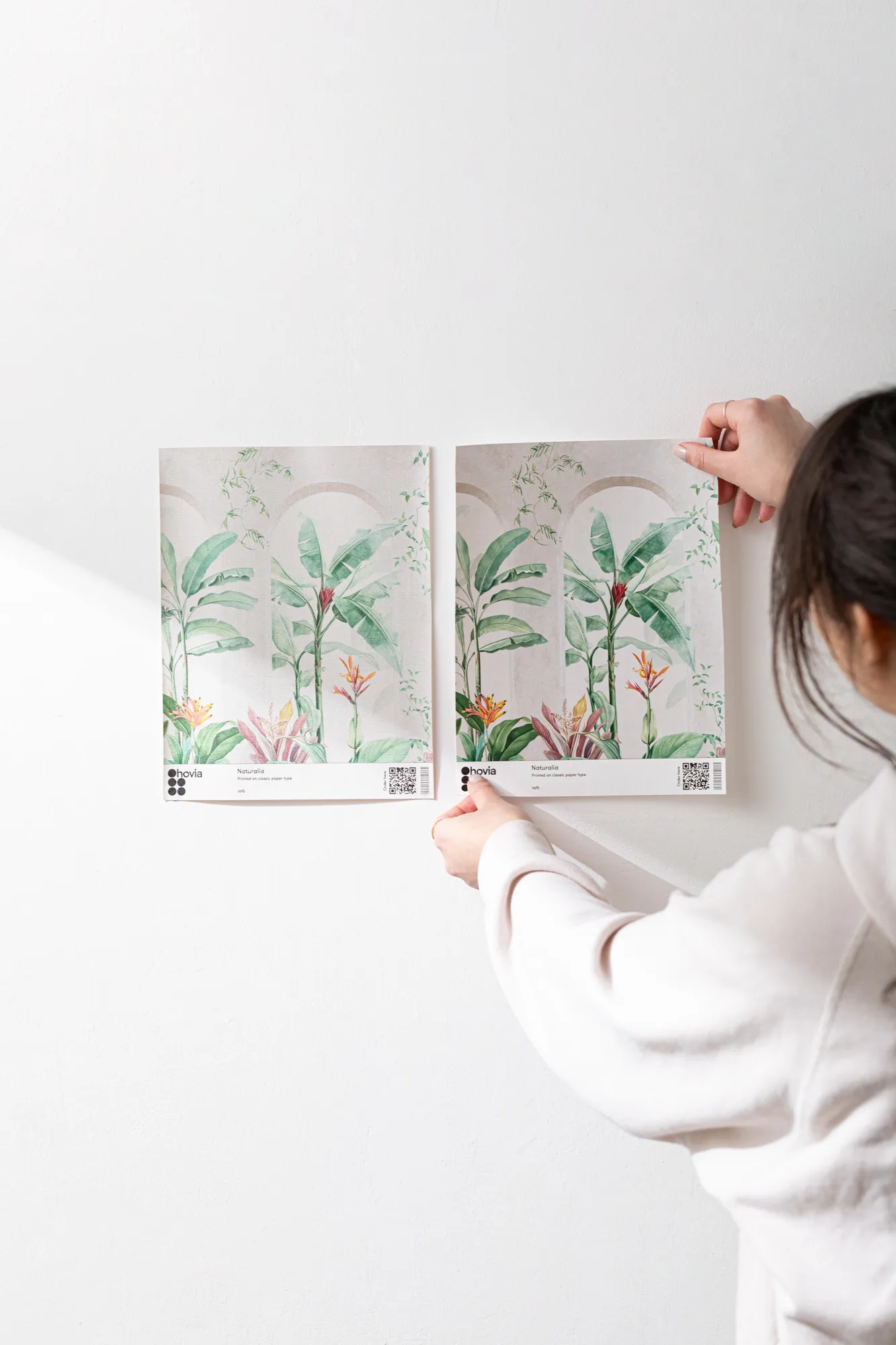 A person holds two tropical wallpaper samples with banana leaves and bird of paradise flowers against a white wall.