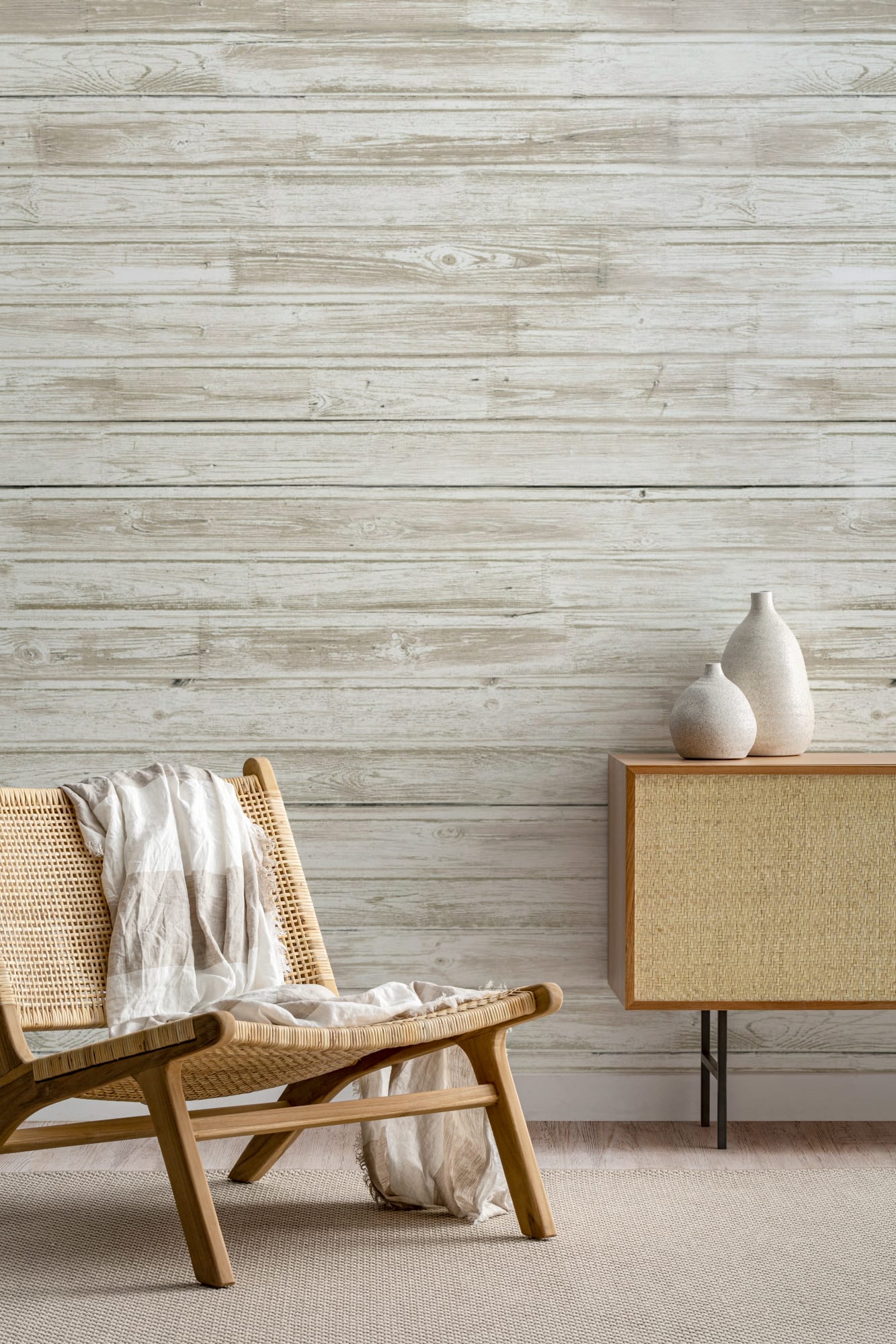 Neutral-toned living room with wood-effect wallpaper, a woven lounge chair draped with a light throw, and a sideboard topped with ceramic vases.