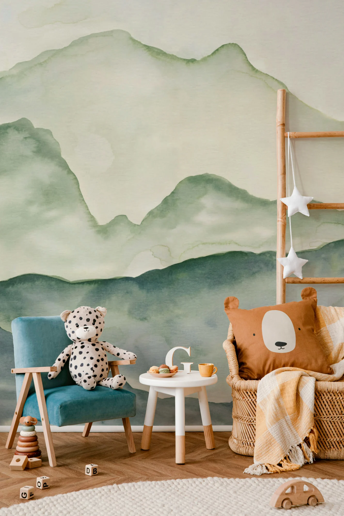 A playful space with a green watercolor mountain wallpaper, teal chair with plush toy, bear cushion, and natural wood toys.