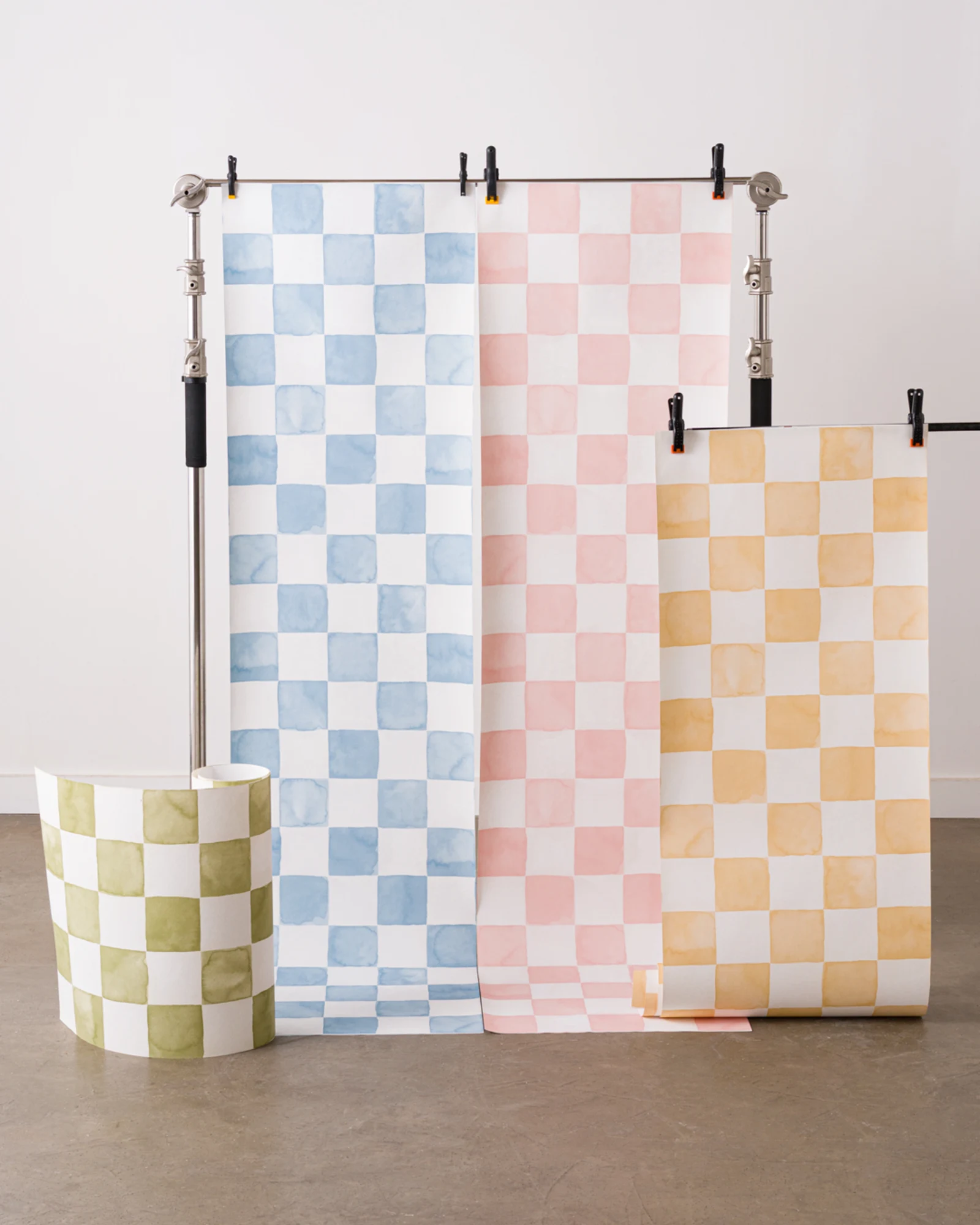 Four panels of watercolor checkered wallpaper in blue, pink, yellow, and green displayed on stands.