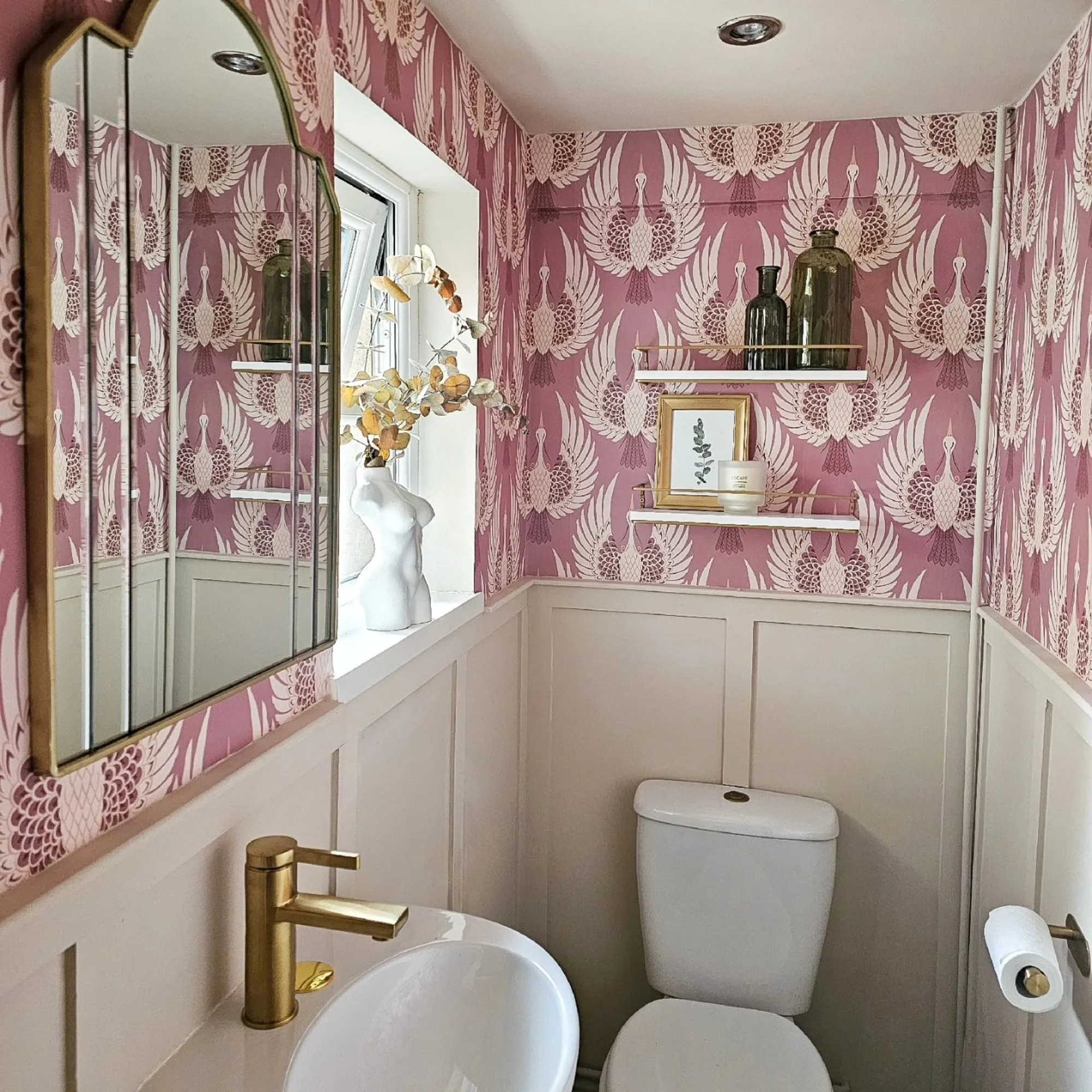 A powder room with pink crane-patterned wallpaper, beige wall paneling, gold fixtures, and a decorative wall shelf.