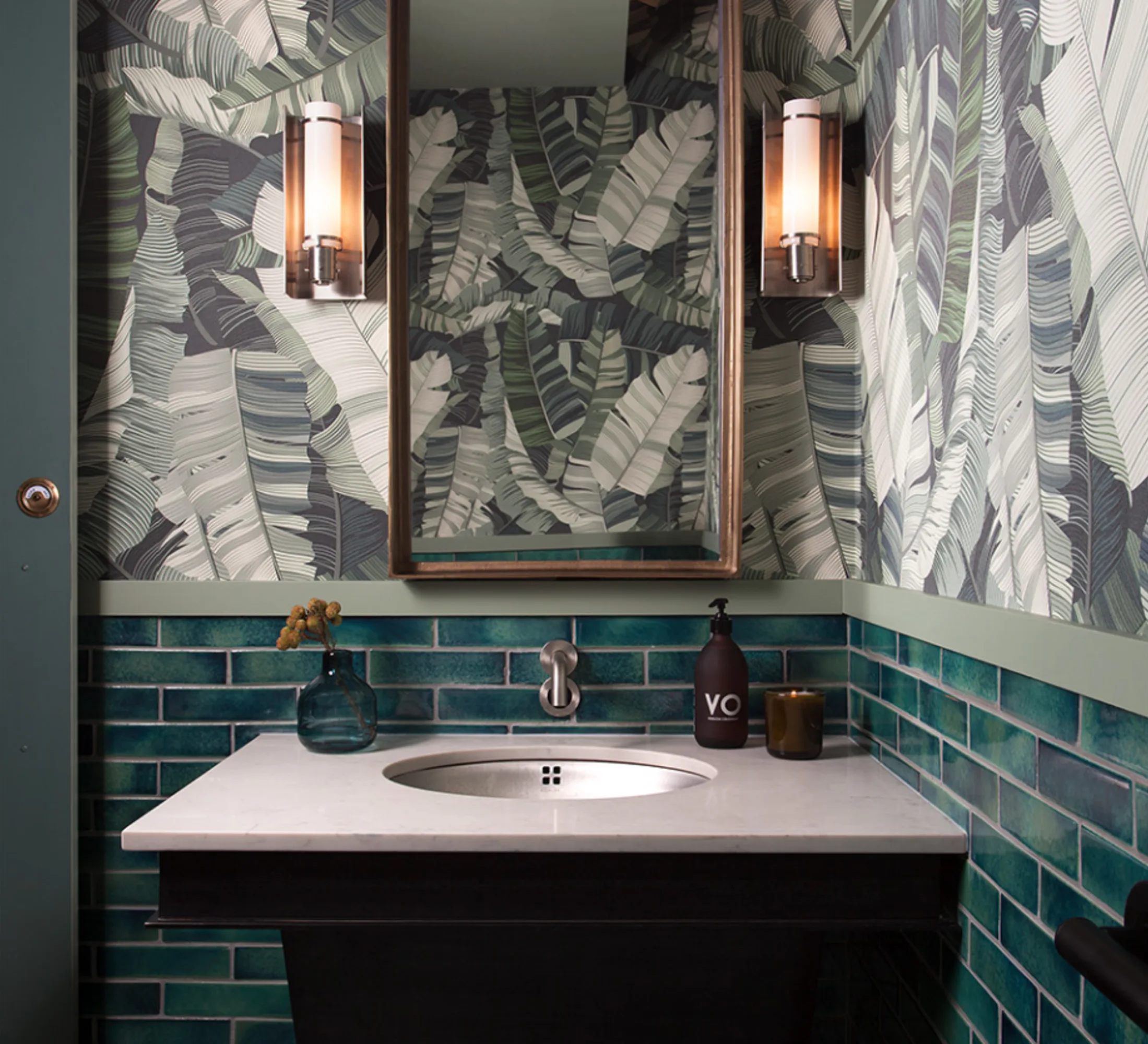 A dark green tropical-themed bathroom with large leafy wallpaper, teal subway tiles, and a marble sink countertop.