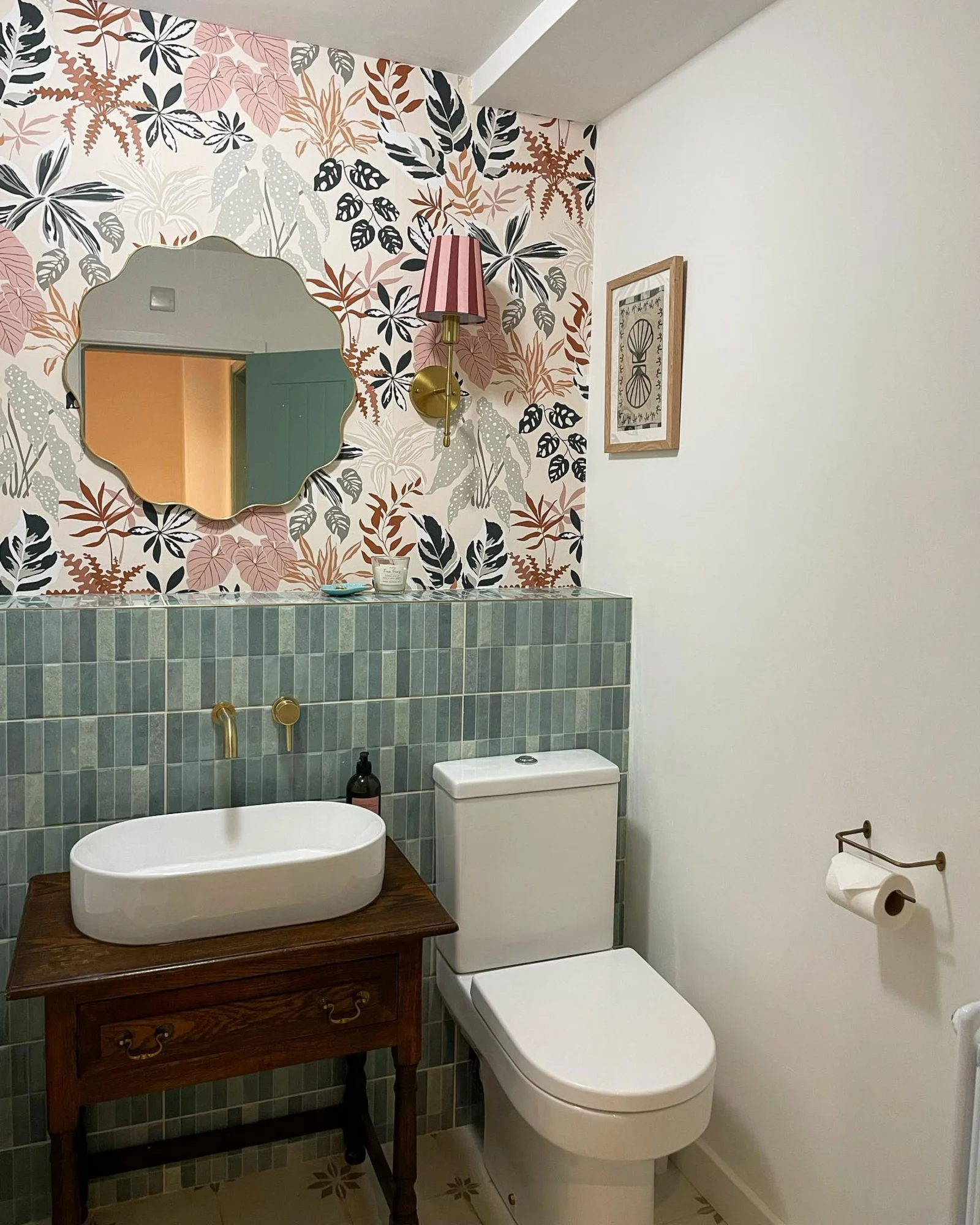A small bathroom featuring botanical wallpaper, green tile backsplash, a white basin sink on a wooden vanity, and a scalloped mirror.