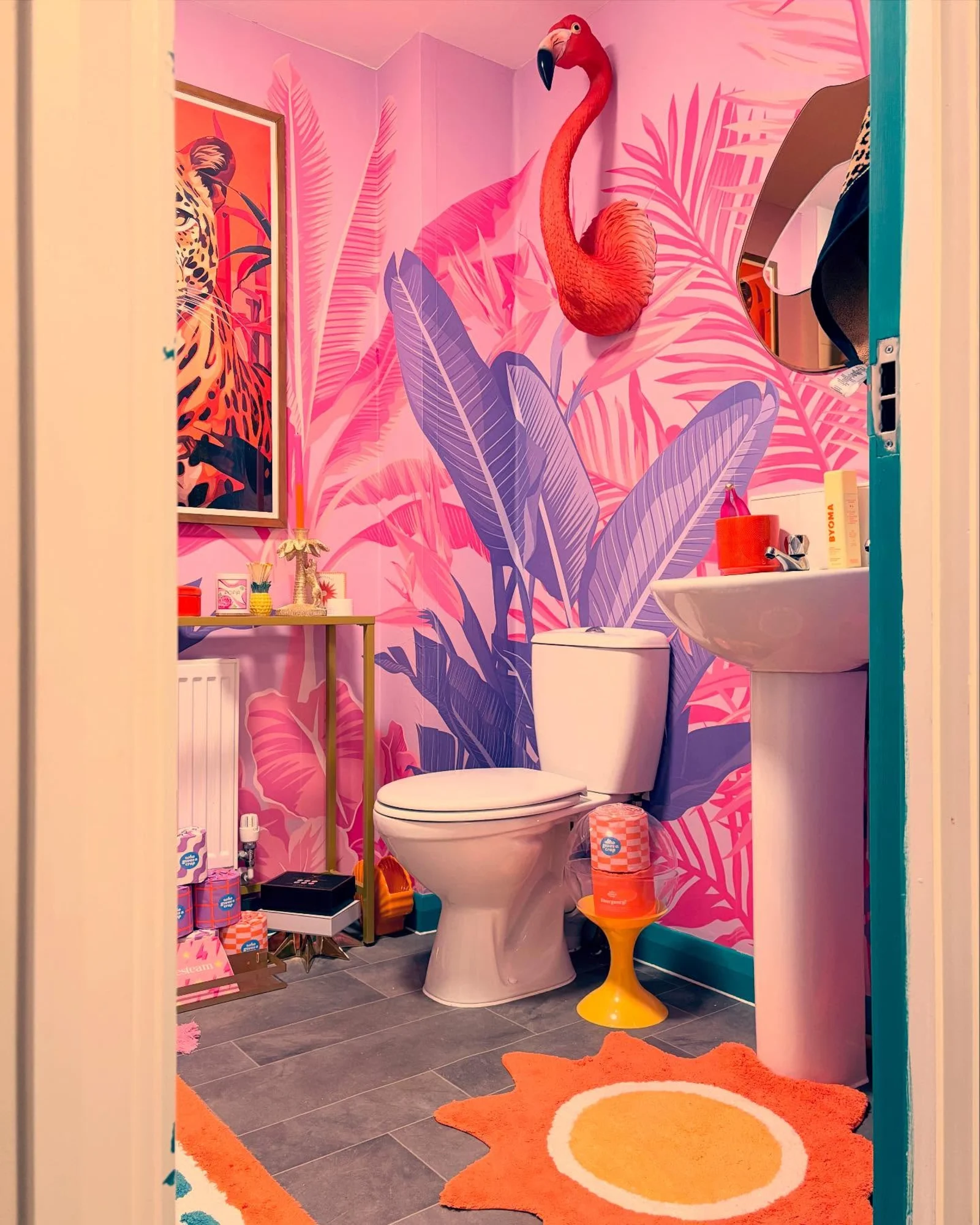 A vibrant bathroom decorated with pink and purple tropical wallpaper, a flamingo wall sculpture, and colorful accessories.