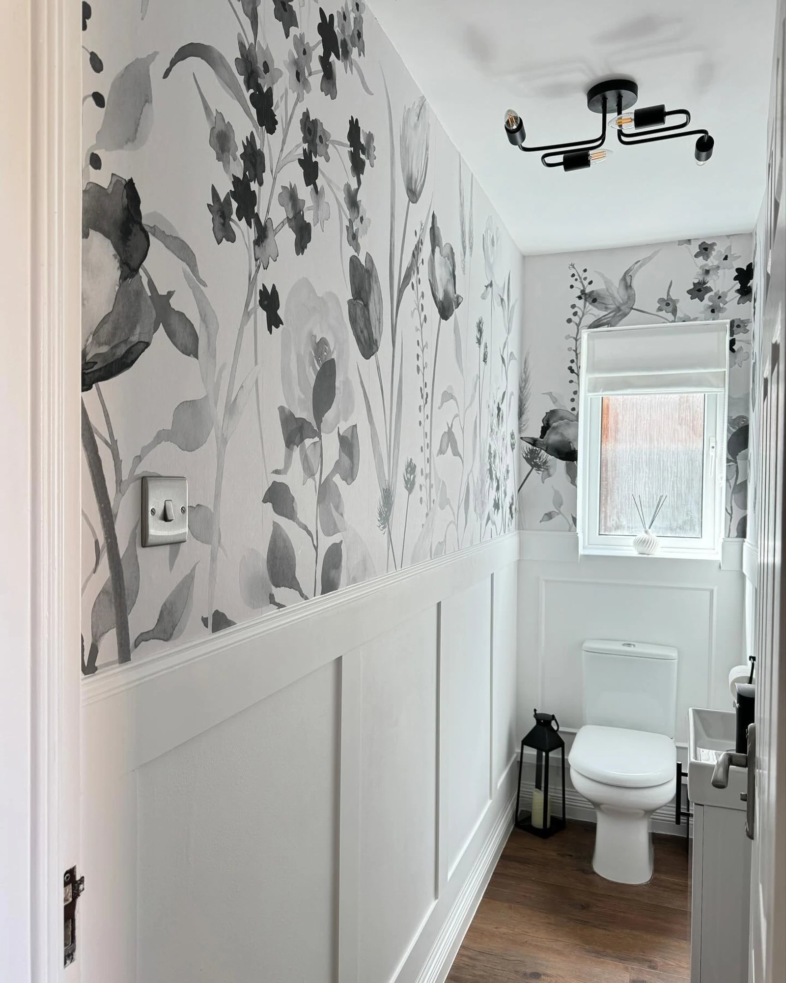 A narrow bathroom with monochrome watercolor floral wallpaper, white wainscoting, and wood-effect flooring.