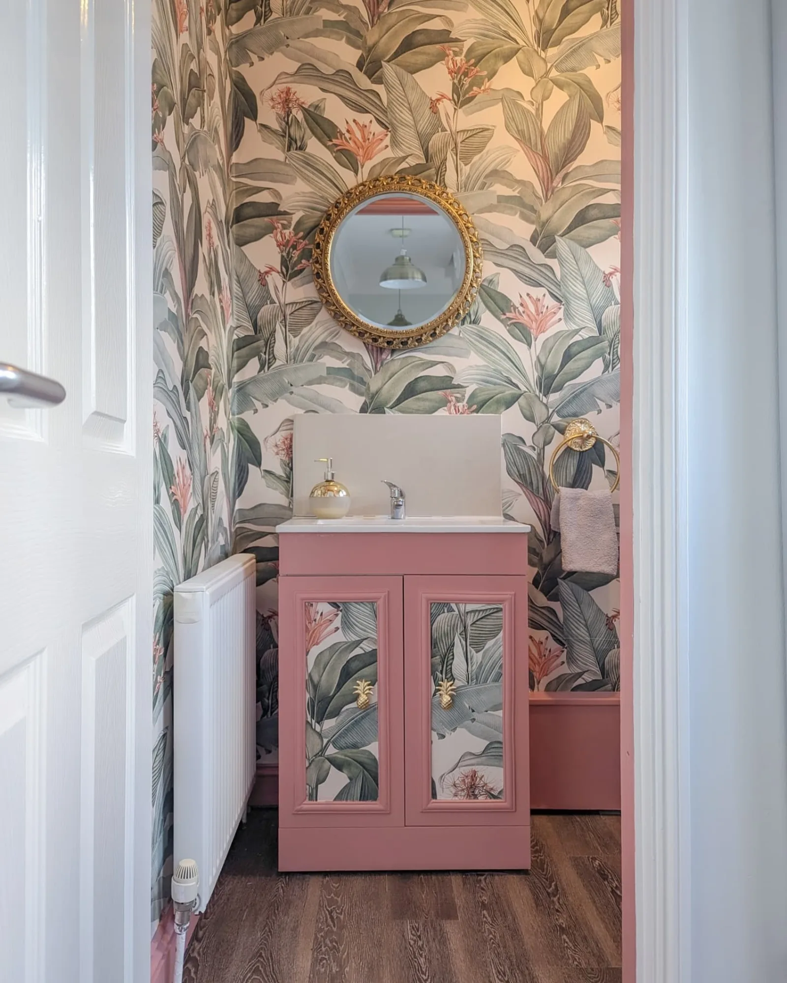 A tropical-inspired powder room with botanical wallpaper, a gold-framed mirror, and a pink vanity cabinet with matching wallpaper.