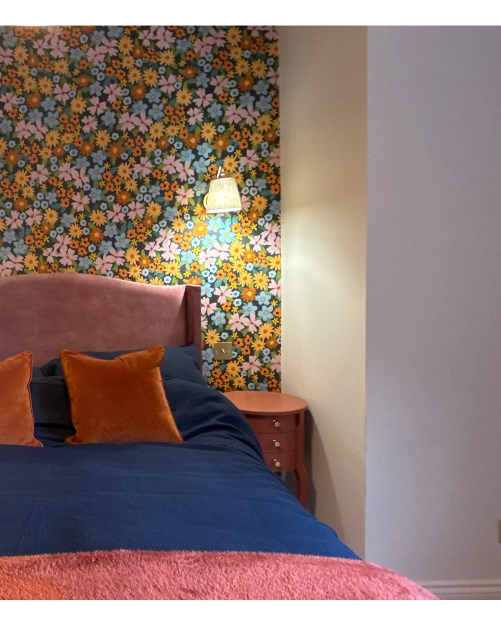 Bedroom with a dark floral wallpaper behind a pink velvet headboard and rust-colored cushions.