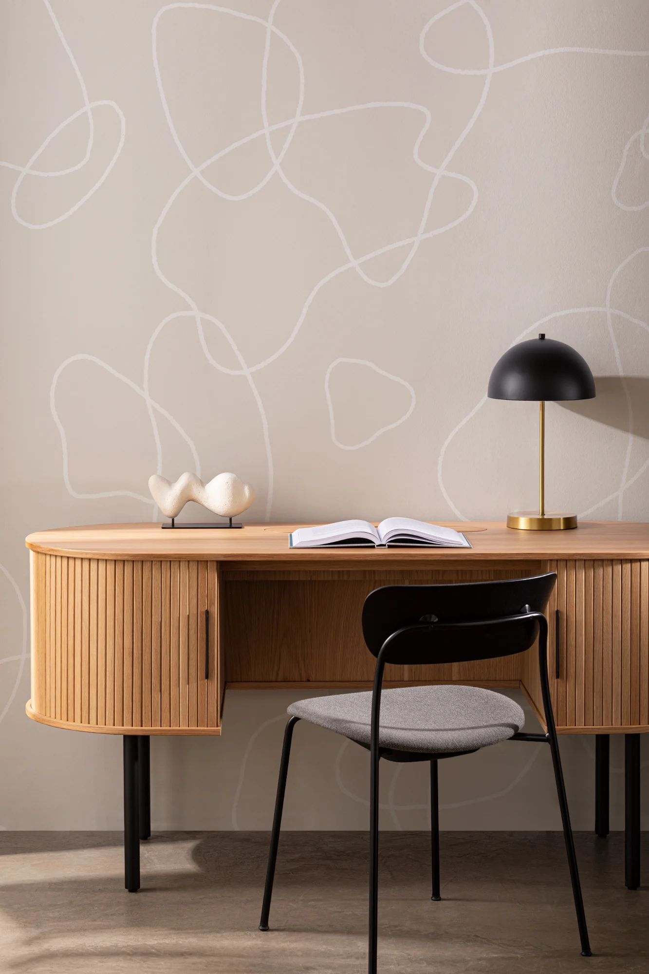 A minimalist home office with a light taupe wall featuring abstract white line art behind a wood desk and black lamp.