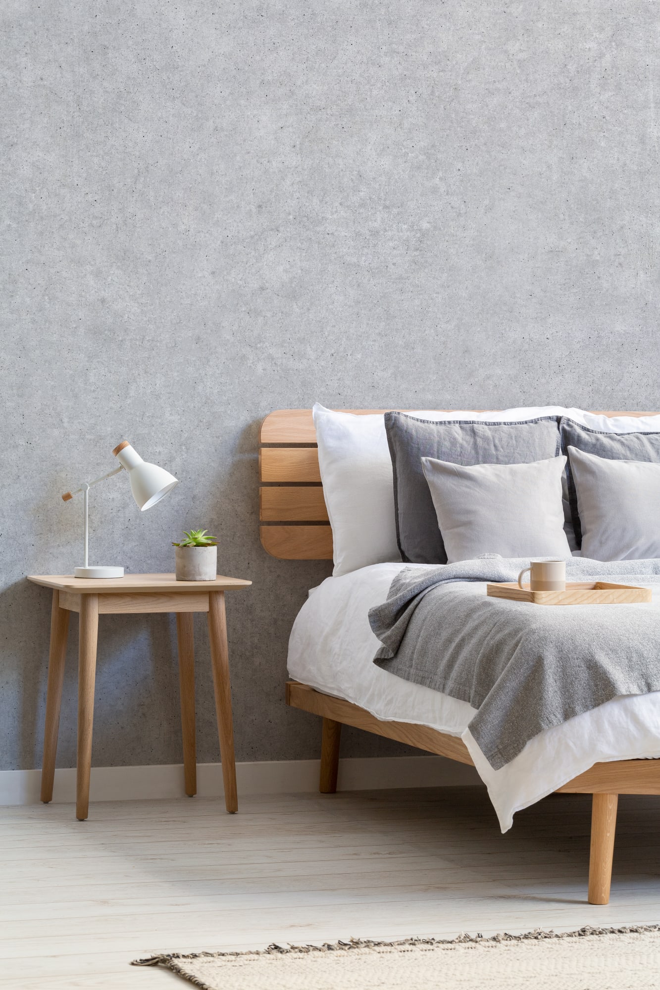 A concrete-look wallpaper in a minimalist bedroom with a wooden bed, white bedding, and gray pillows.