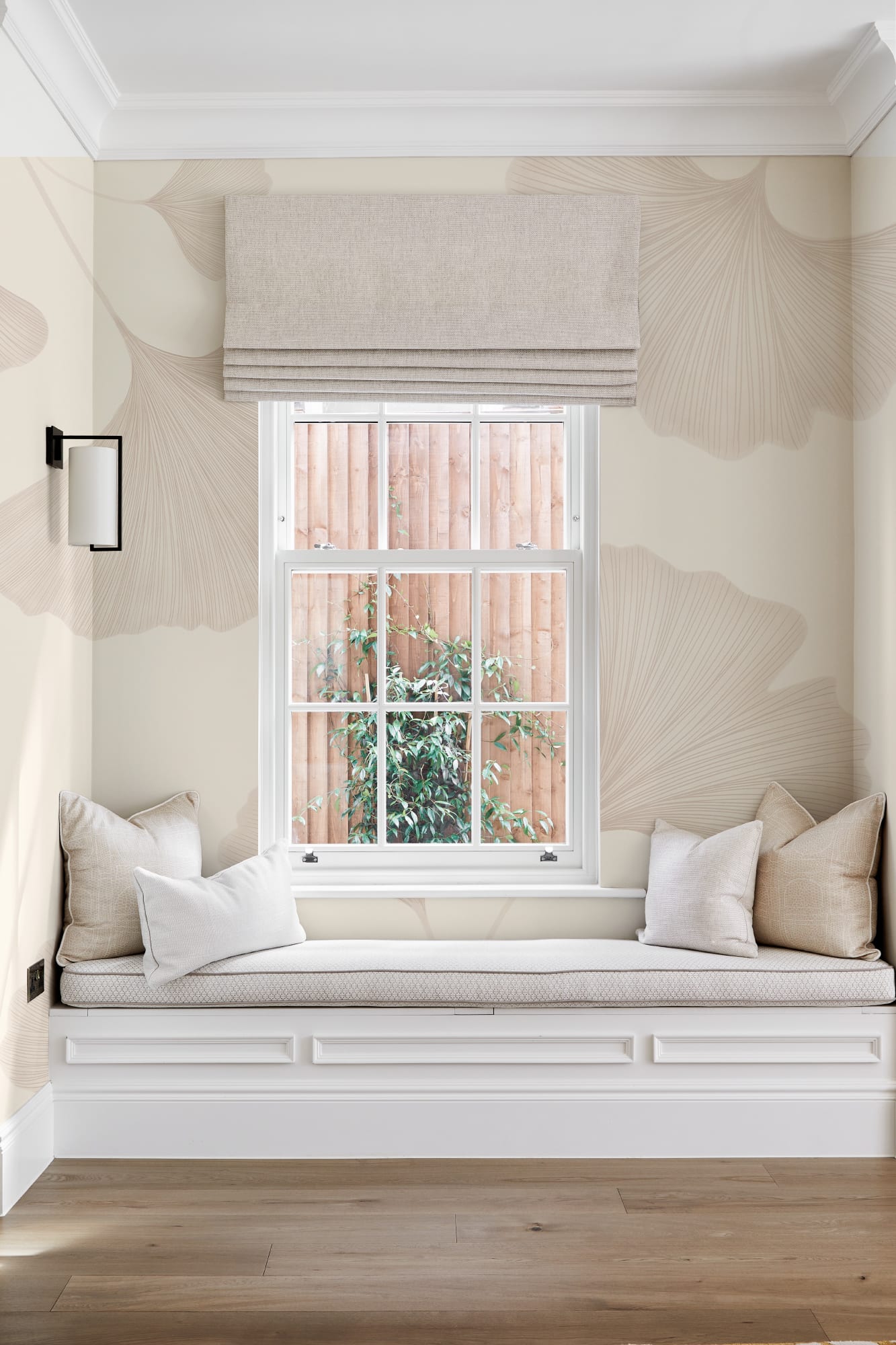 Neutral-toned room with a window seat, cream floral wallpaper, and beige cushions surrounding a white-framed window.