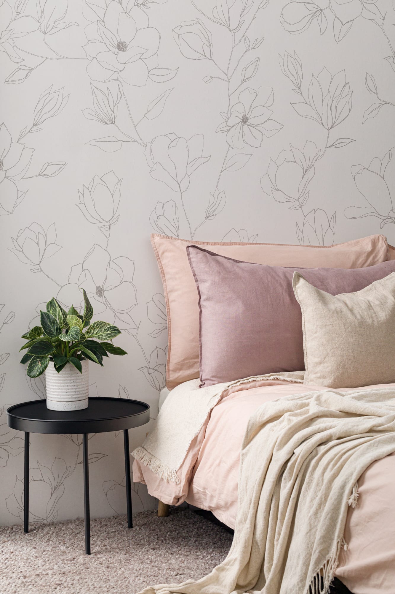 Close-up of a bedroom with soft pink bedding and a white floral line-art wallpaper behind a black side table with a potted plant.