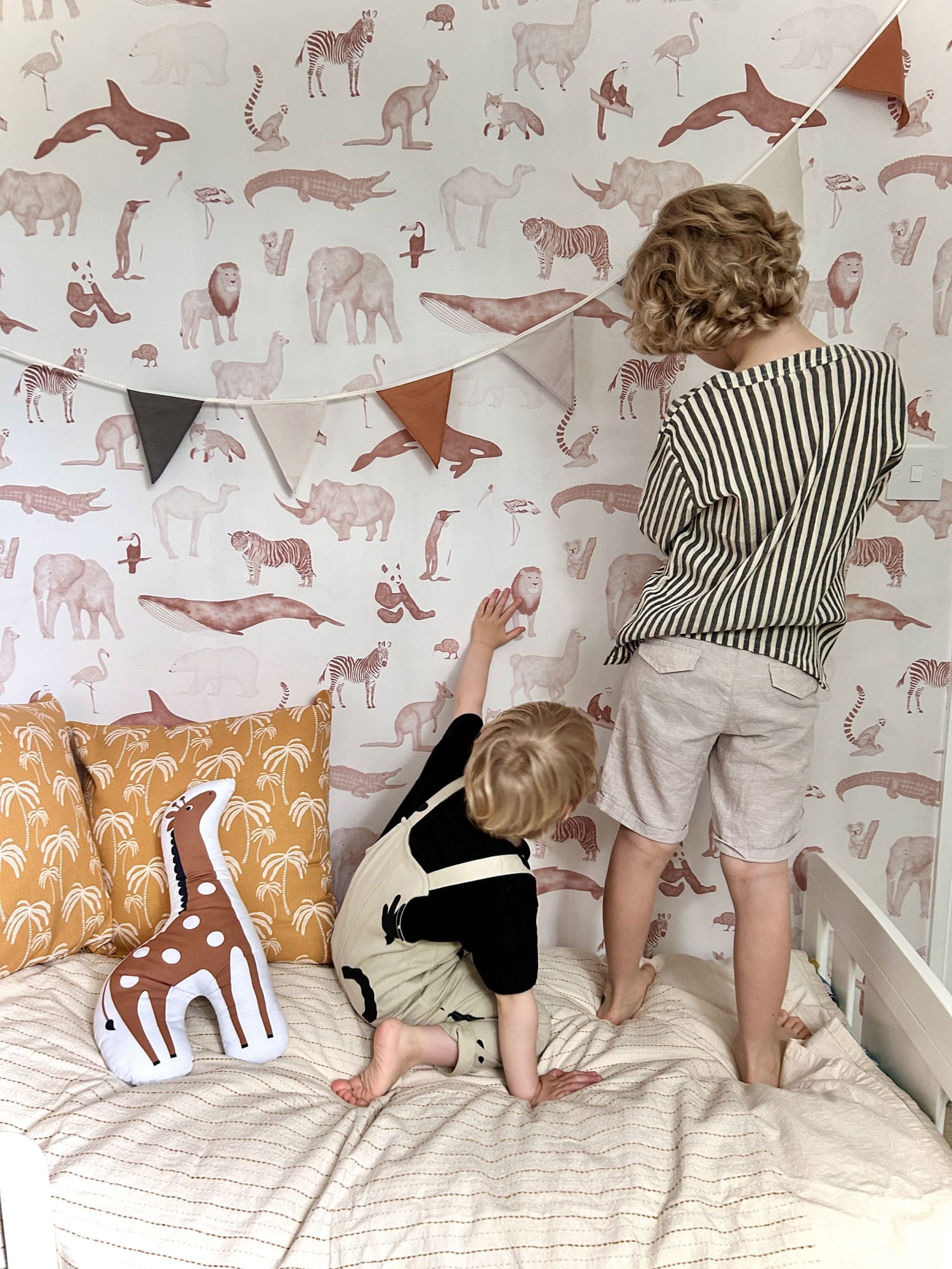 Two children play on a bed against a watercolor animal-print wallpaper in soft terracotta tones.