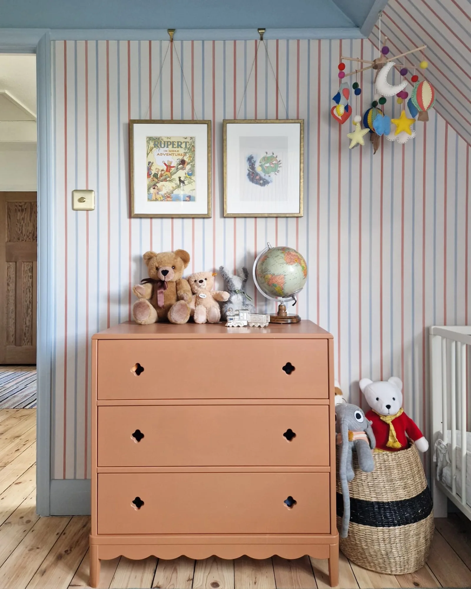 Striped wallpaper with a terracotta dresser, teddy bears, framed artwork, and a colorful hanging mobile in a cozy nursery corner.