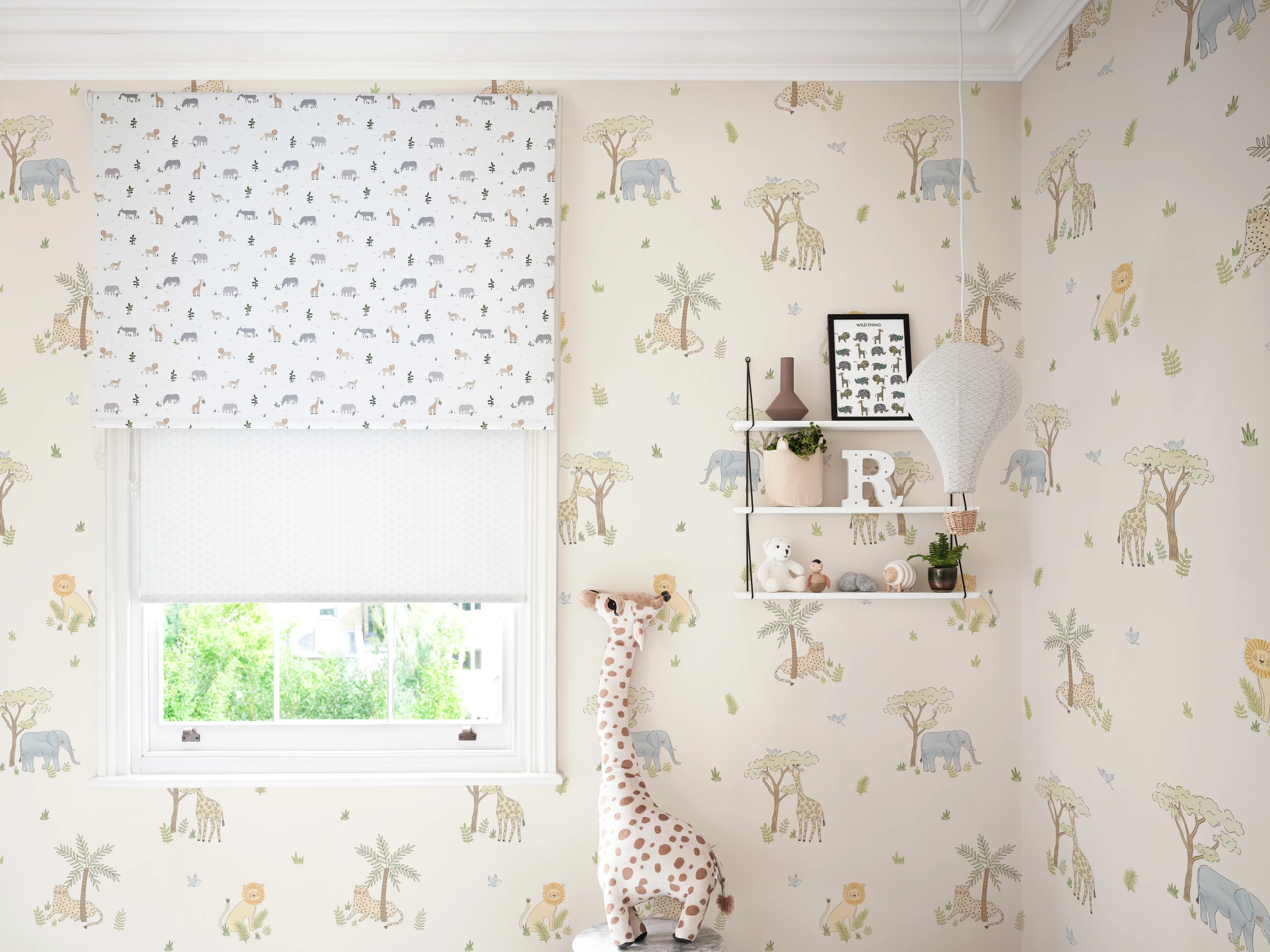 Safari-themed nursery features animal and tree wallpaper with a toy giraffe, white shelves, and a hot air balloon light.