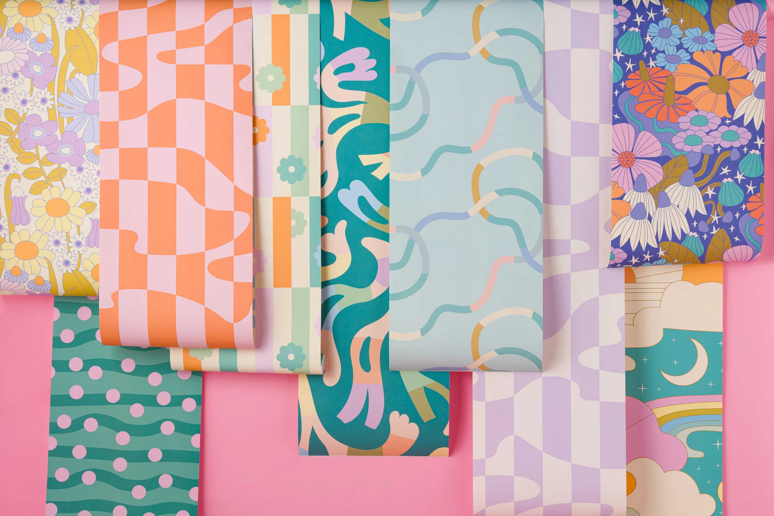 Assorted colorful wallpaper rolls with playful retro and floral patterns arranged on a pink surface.