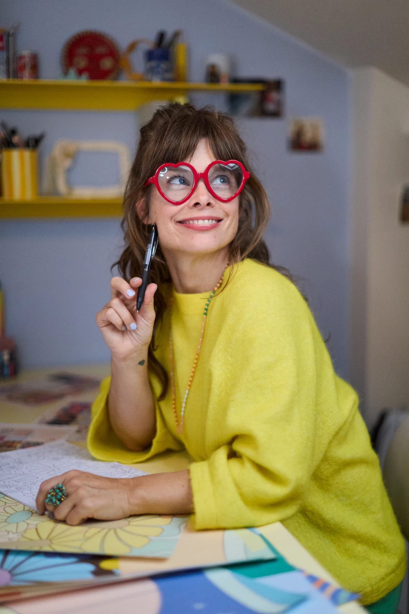 Elizabeth Olwen wearing red heart-shaped glasses and a yellow sweater smiles while holding a pen at a creative desk.
