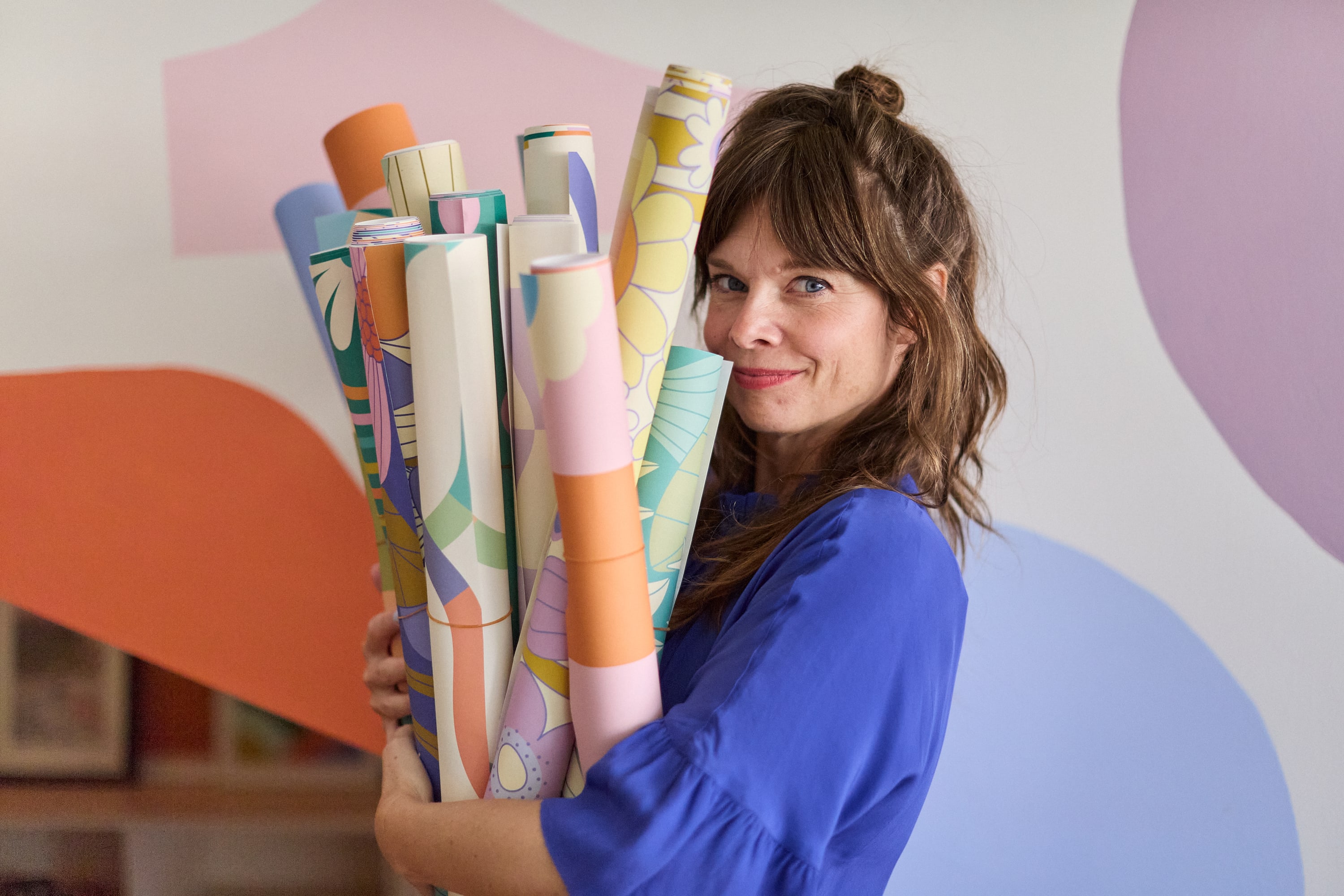 Elizabeth Olwen in a blue dress smiles while holding colorful rolled-up wallpaper designs.