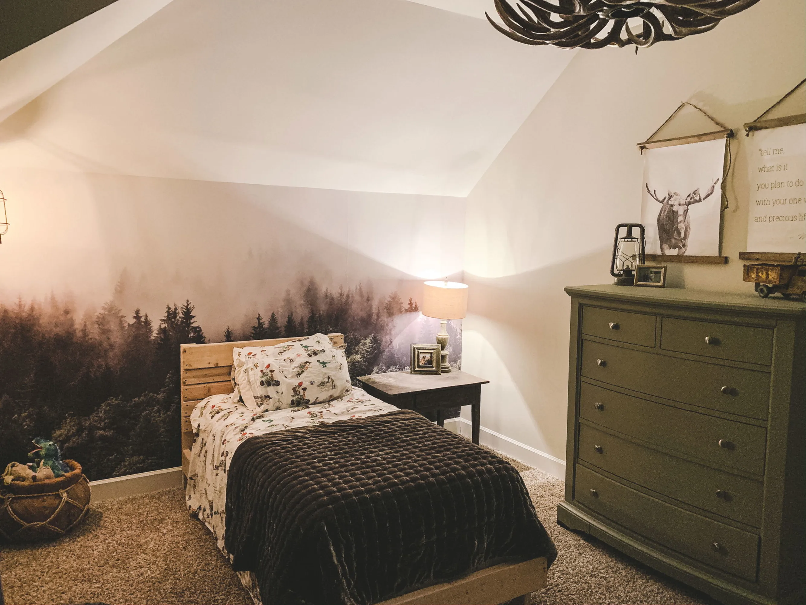 Cozy bedroom with forest mural wallpaper behind a child’s bed under a sloped ceiling.