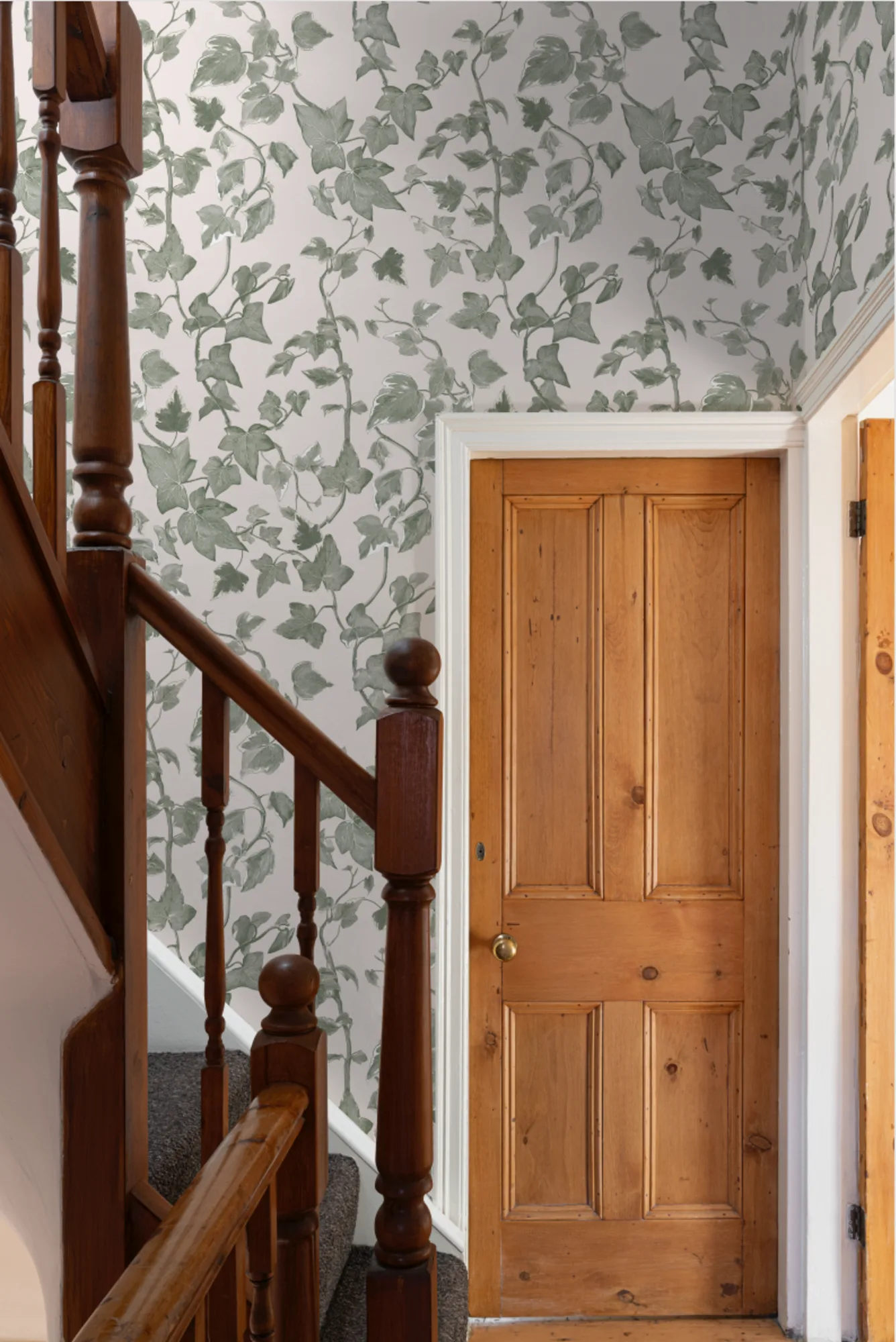 A stairway hallway features pale green leafy wallpaper with a natural wood door and bannisters.