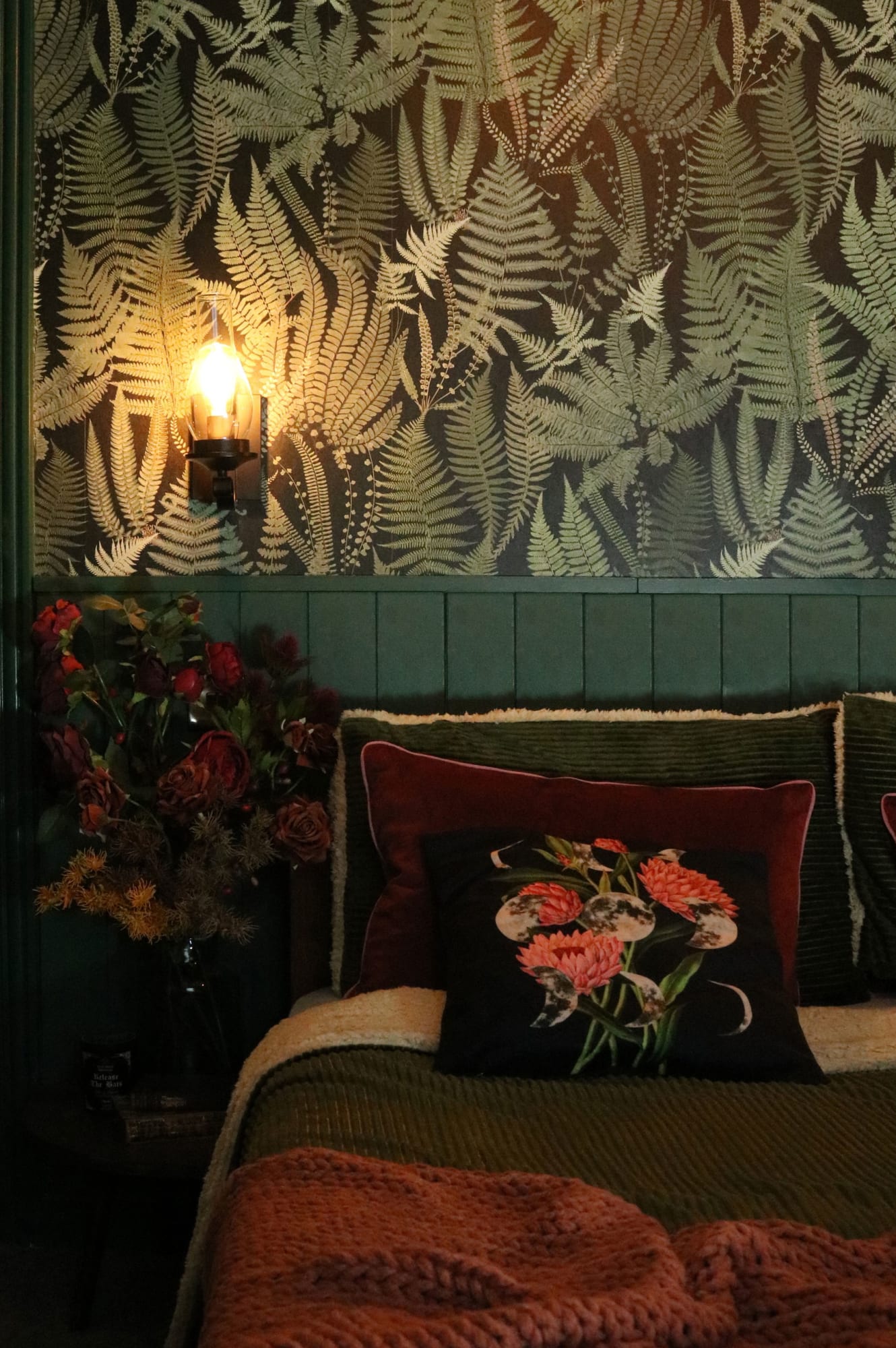 A dark green bedroom with a fern wallpaper and cozy layered textiles.
