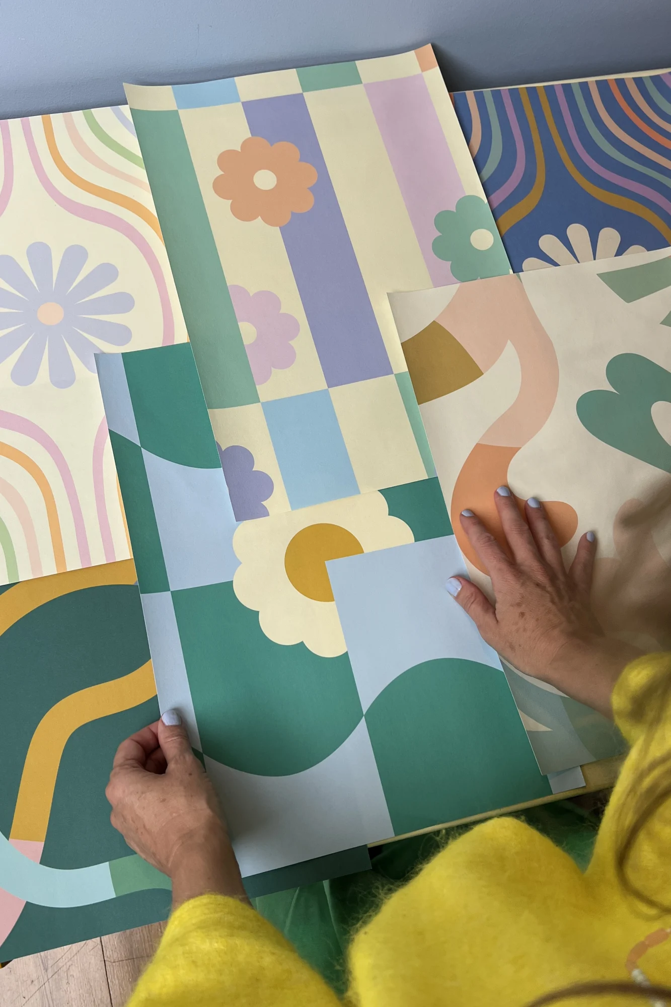 Elizabeth Olwen in a yellow sweater places retro-style wallpaper samples on a table with a blue wall.