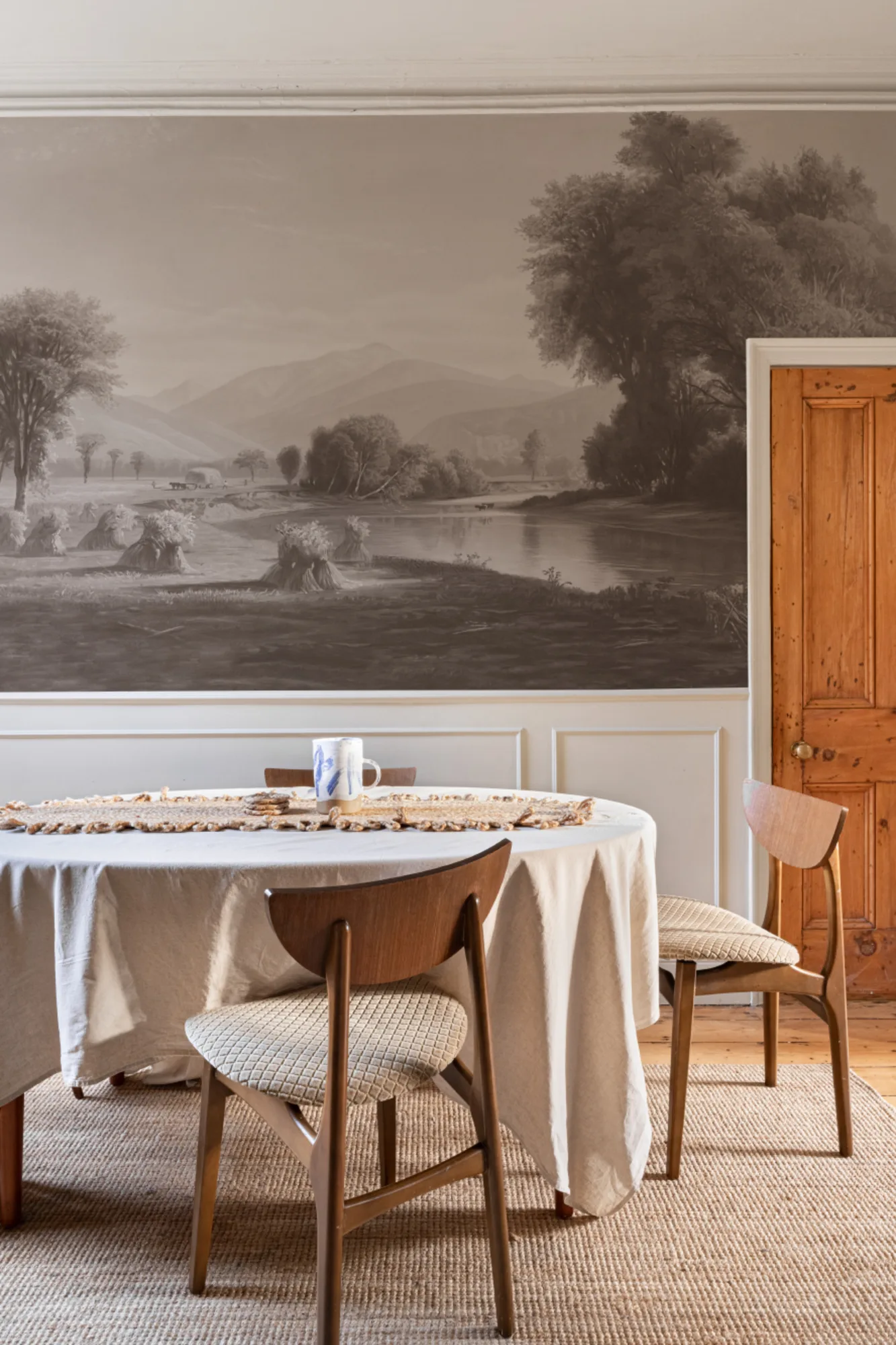 A detailed grayscale countryside mural decorates the walls of a dining room with a round table and vintage chairs