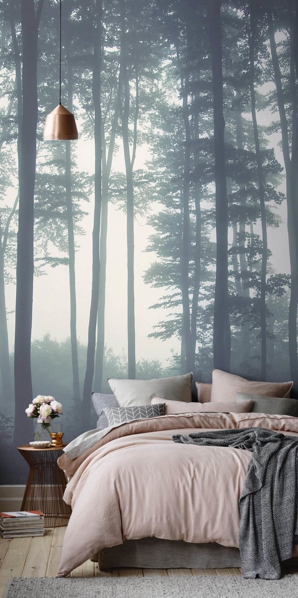 Bedroom featuring a wall mural of a misty forest, with a bed dressed in pink bedding, surrounded by gray and patterned pillows. A round wooden side table displays pink flowers, enhanced by a copper pendant light above.